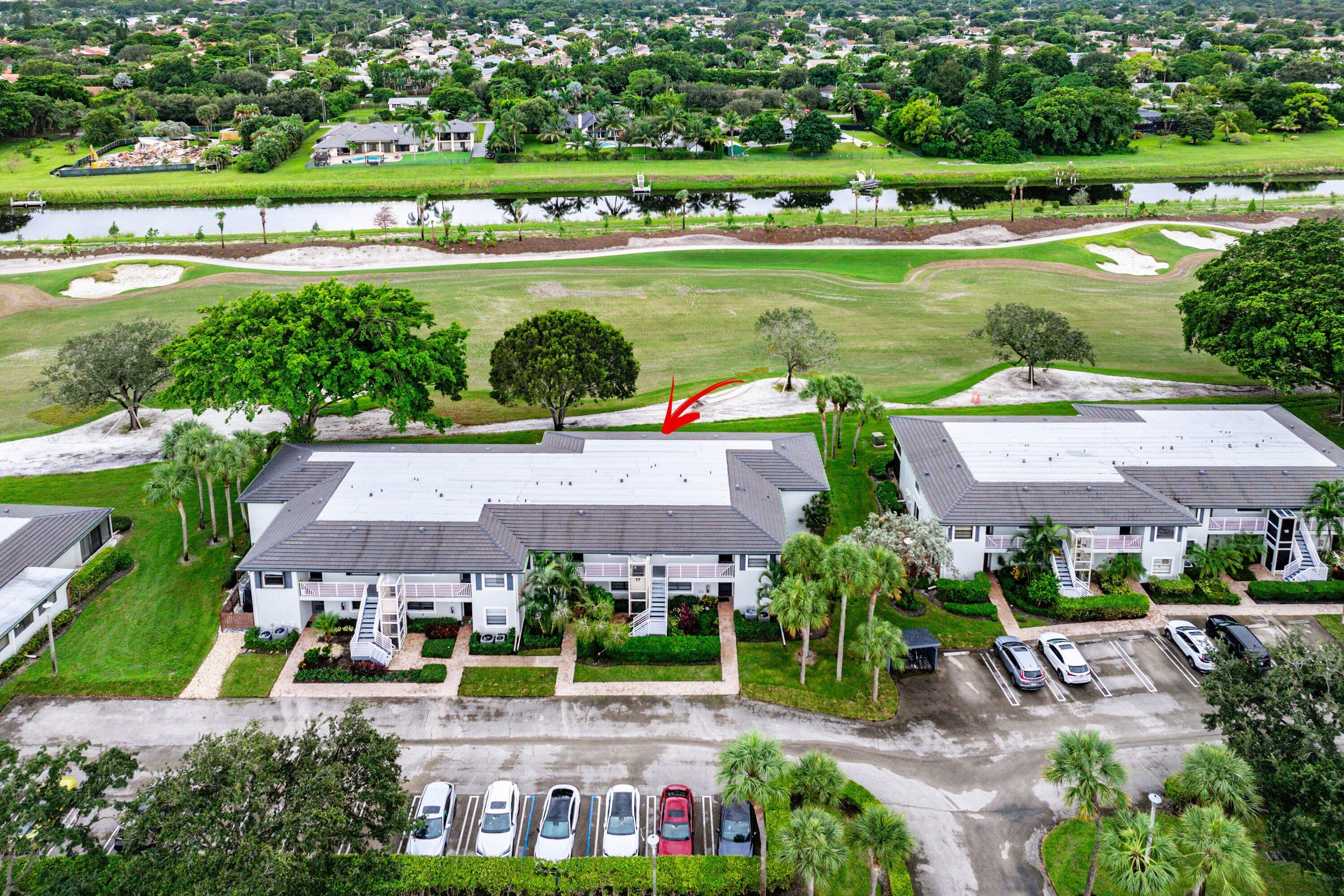 Aerial perspective of a multi-unit residential building featuring a multi-hipped roof, paved parking area, landscaping, and immediate proximity to a golf course and canal