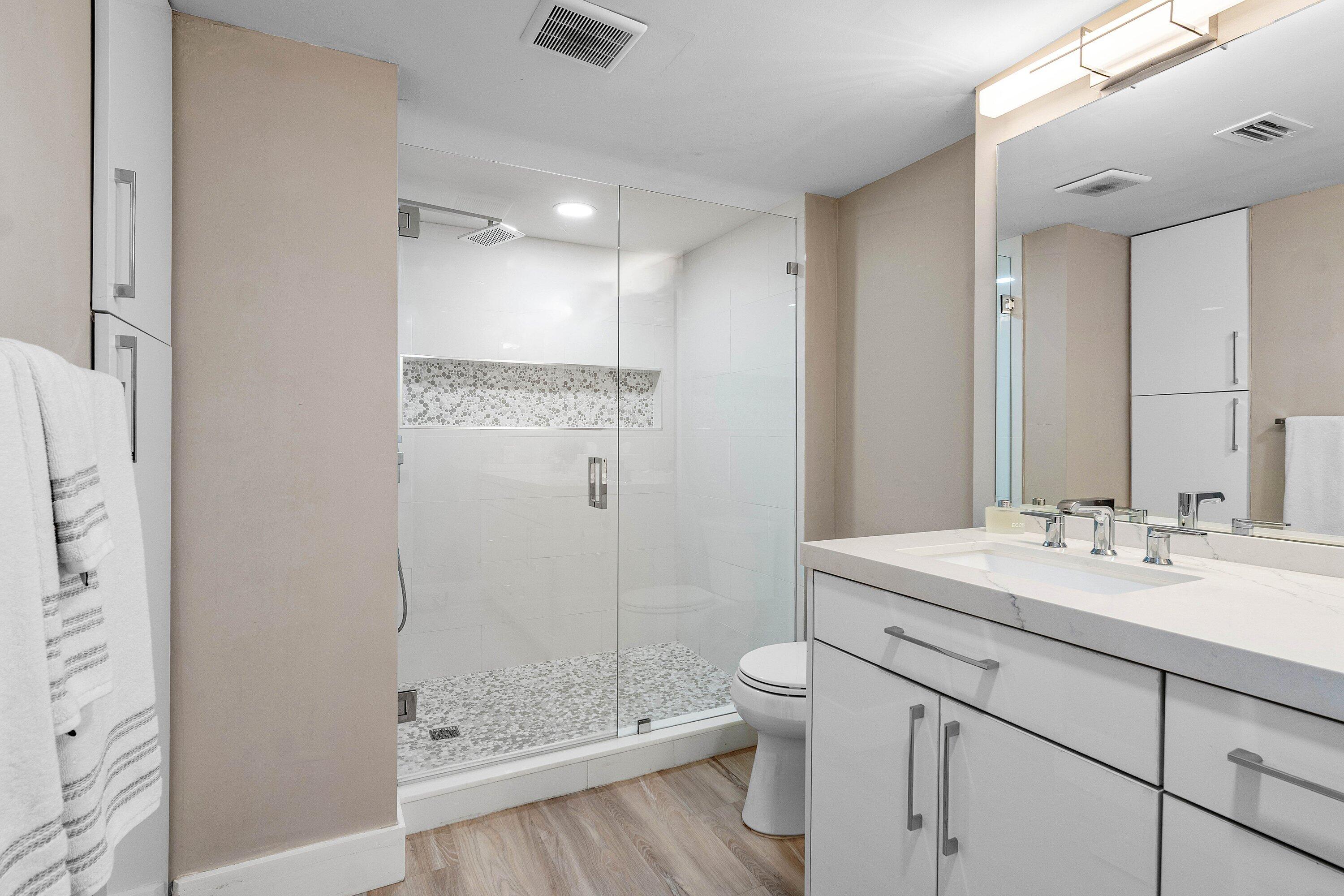 Bathroom featuring a white vanity with integrated rectangular sink and quartz countertop, glass-enclosed shower with tiled pebble-style flooring and built-in niche, and wood-finish flooring
