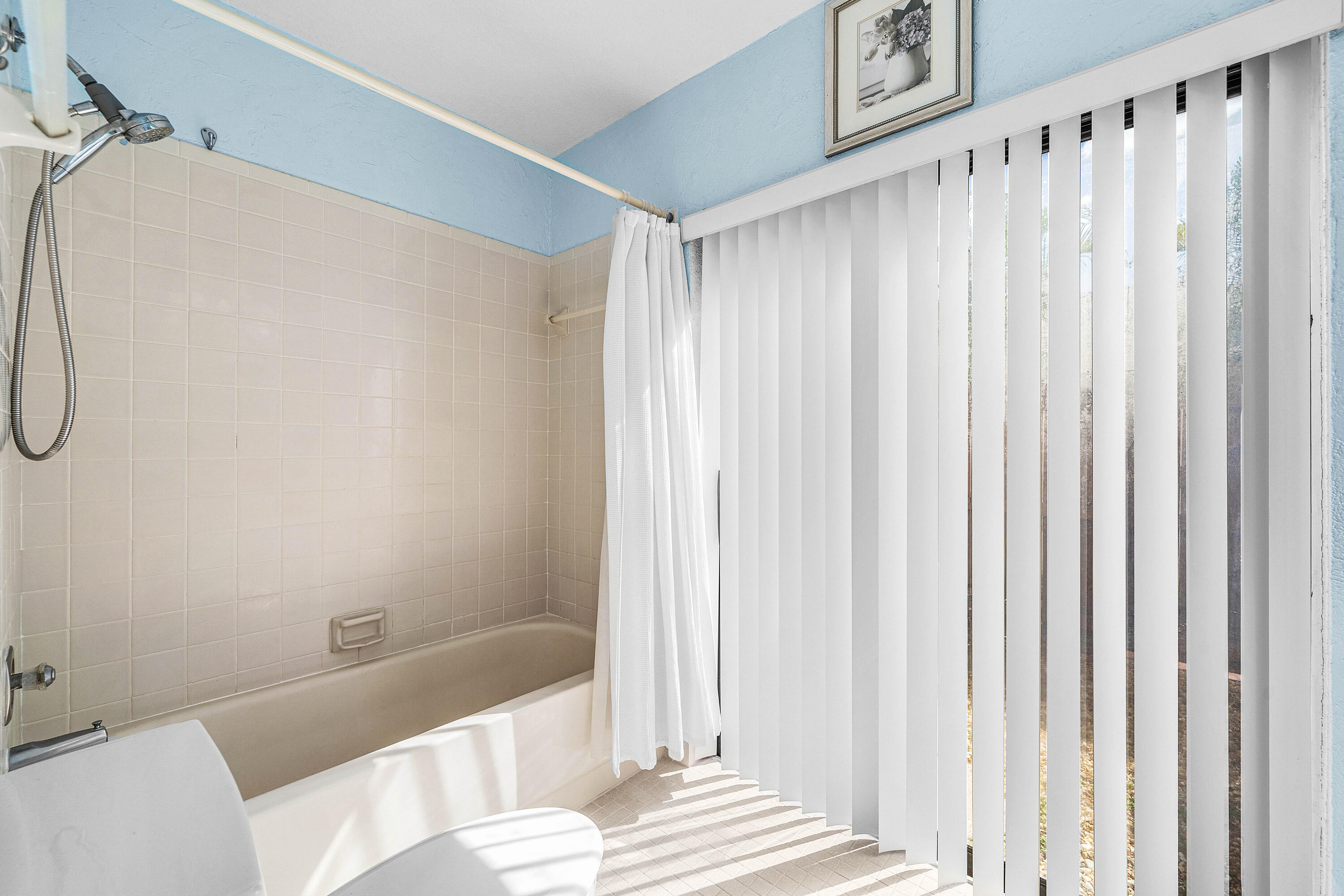 Bathtub enclosure with tiled wall surround, wall-mounted hand-held shower fixture, and floor-to-ceiling vertical blinds