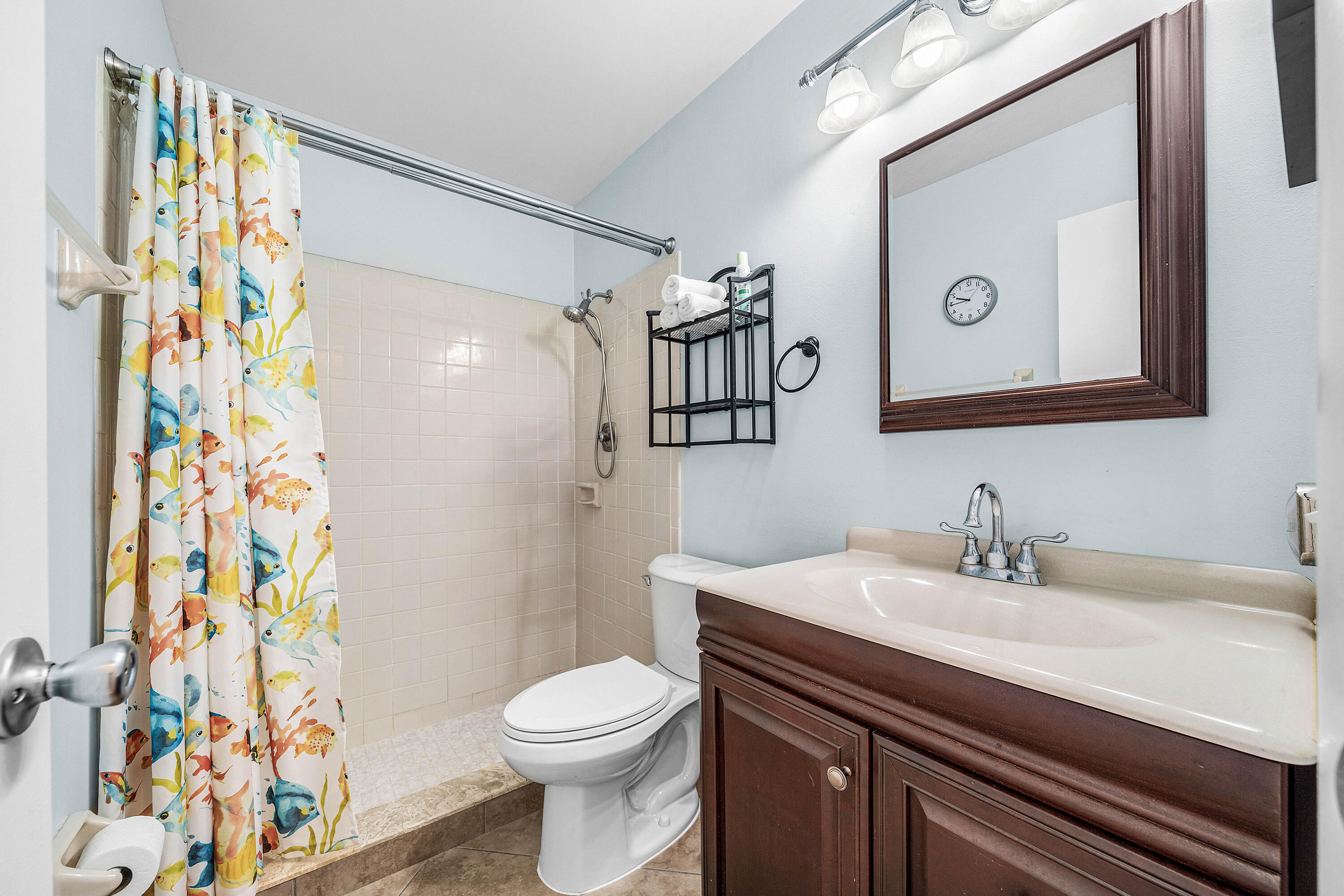 Bathroom featuring a vanity with an integrated countertop, dark wood-finish cabinetry, a framed wall mirror, a multi-light fixture, and a tiled shower stall with a handheld showerhead