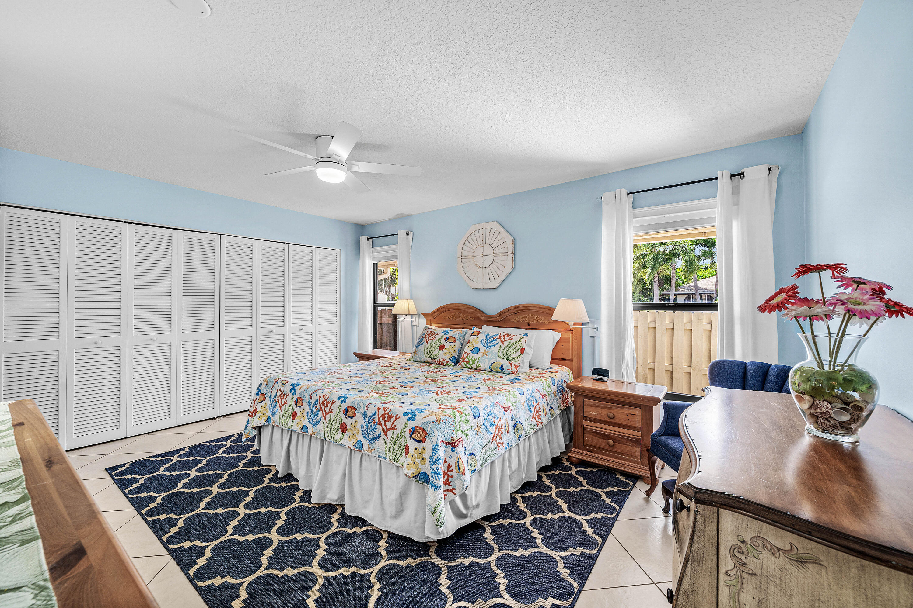 Spacious room featuring light-toned tile flooring, large louvered-style built-in closet doors, a white ceiling fan with integrated lighting, and dual windows with window treatments
