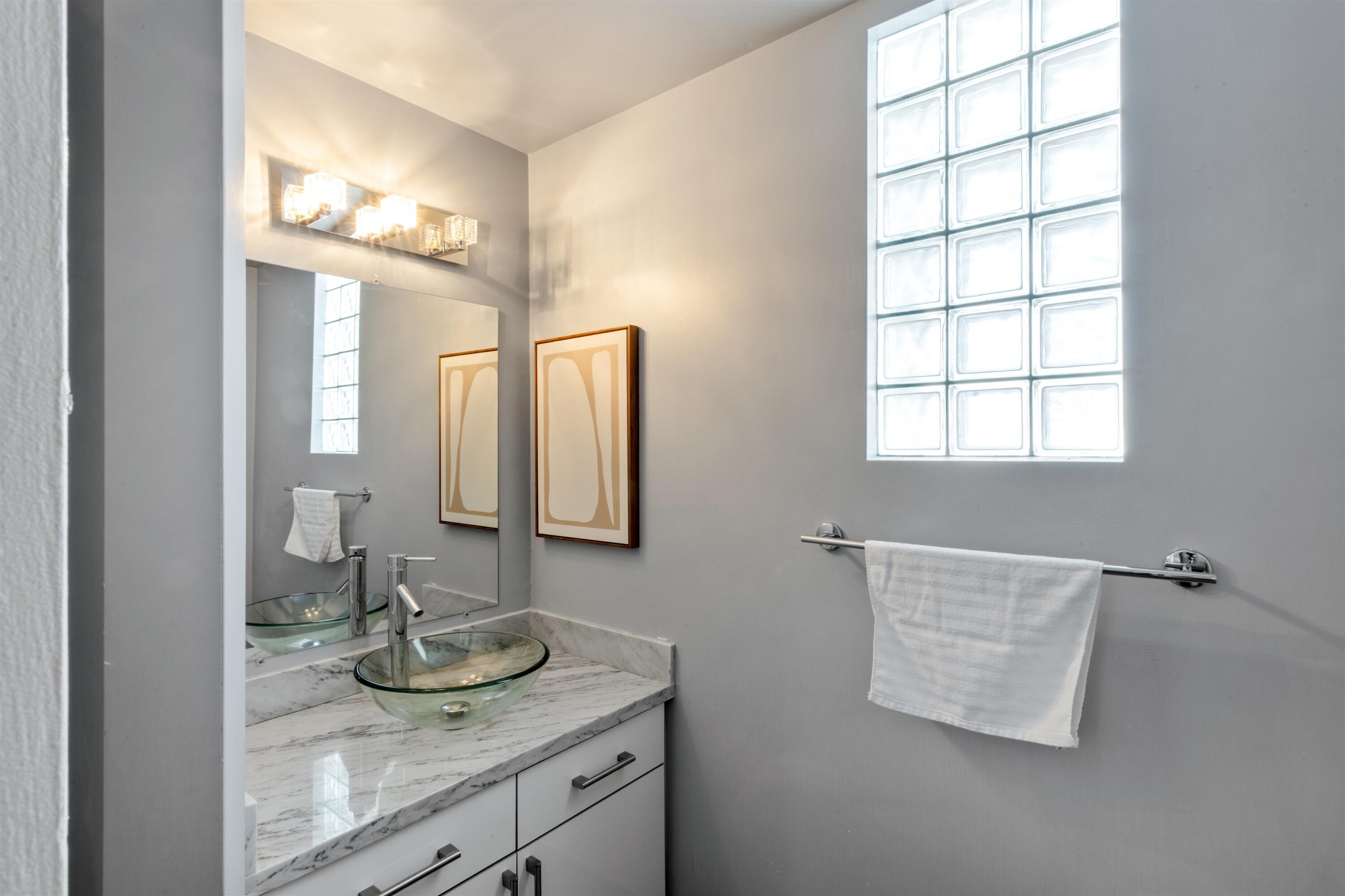Stone-finish countertop featuring a glass vessel sink, modern chrome faucet, built-in cabinetry, and a glass block window