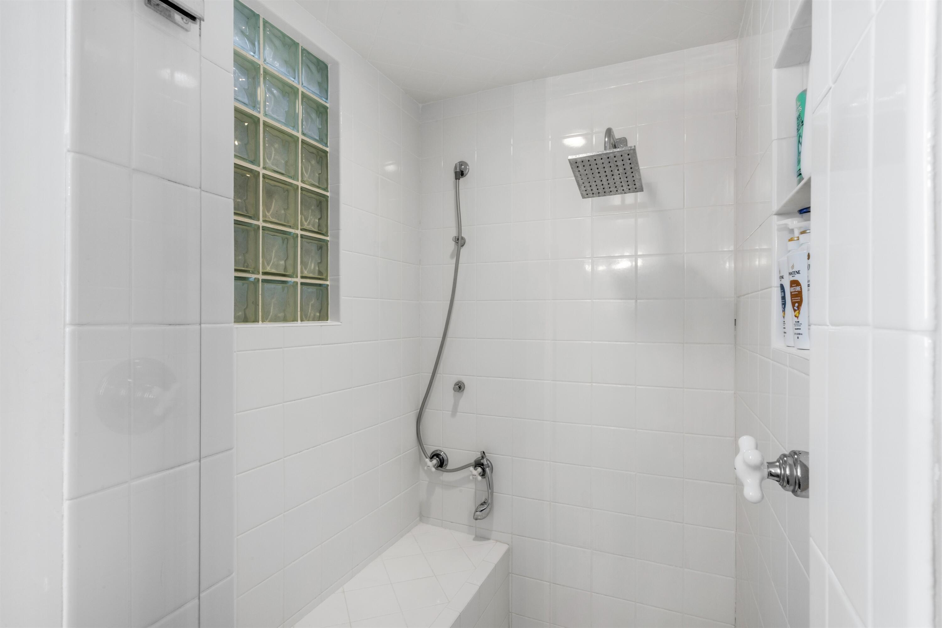 Walk-in shower featuring white tile surround, glass block accent window, rainfall showerhead with handheld attachment, built-in bench seating, and recessed storage niche
