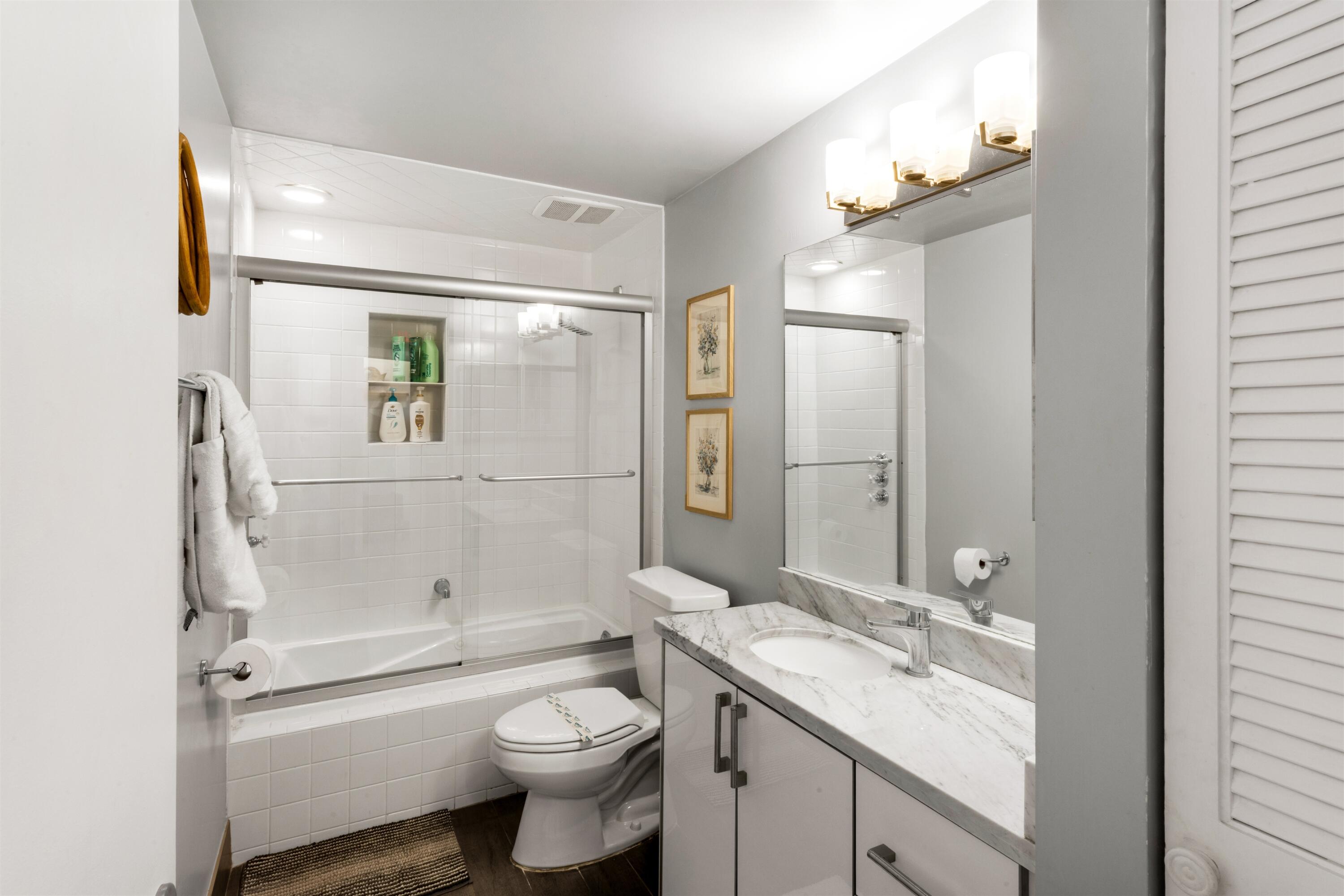 Bathroom featuring a vanity with a stone-top counter, undermount sink, and chrome fixtures, paired with a shower-tub combination featuring a sliding glass enclosure and built-in tiled niche