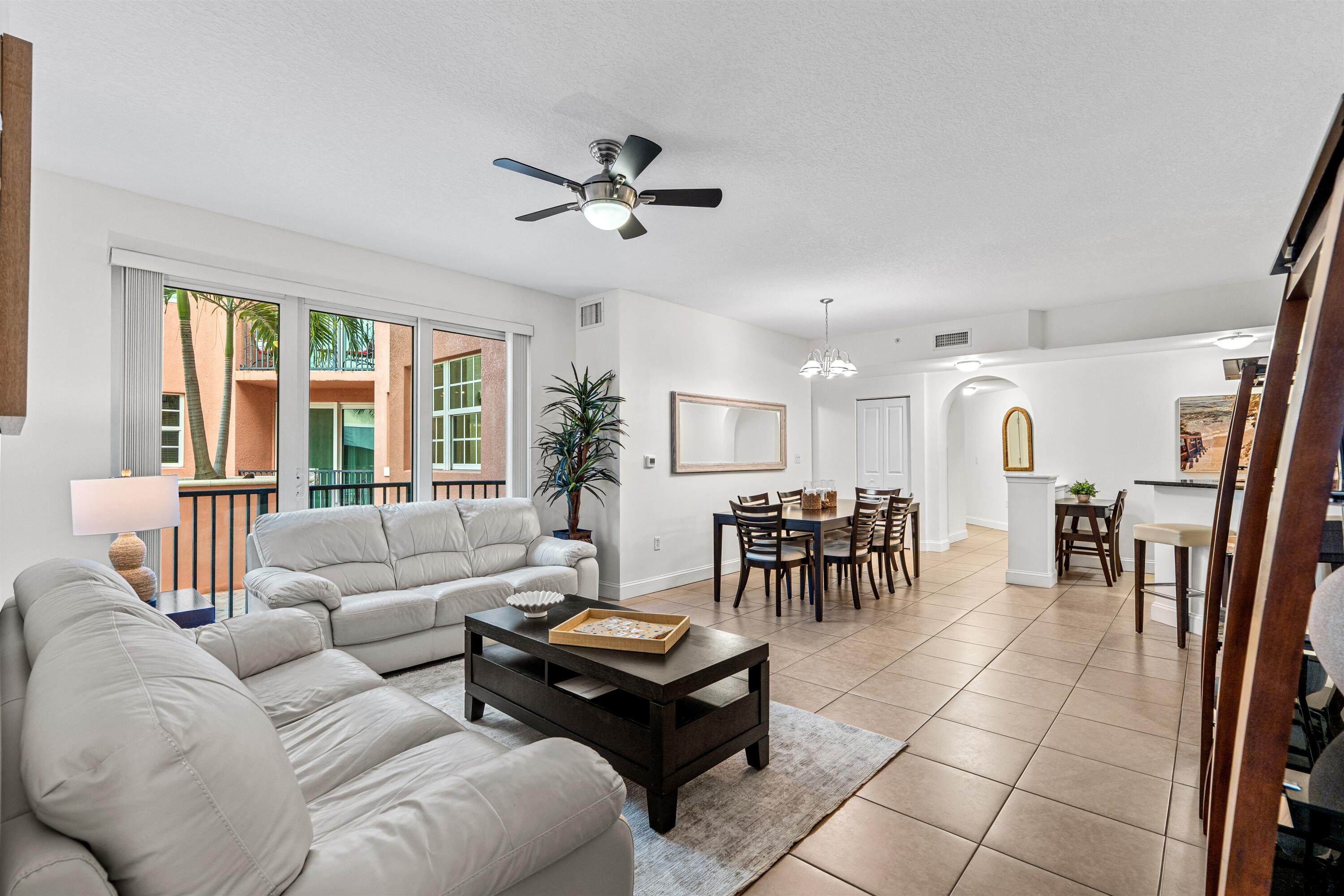 Open-concept interior featuring large-format ceramic tile flooring, sliding glass doors to a balcony, a ceiling fan with integrated lighting, and a kitchen breakfast bar