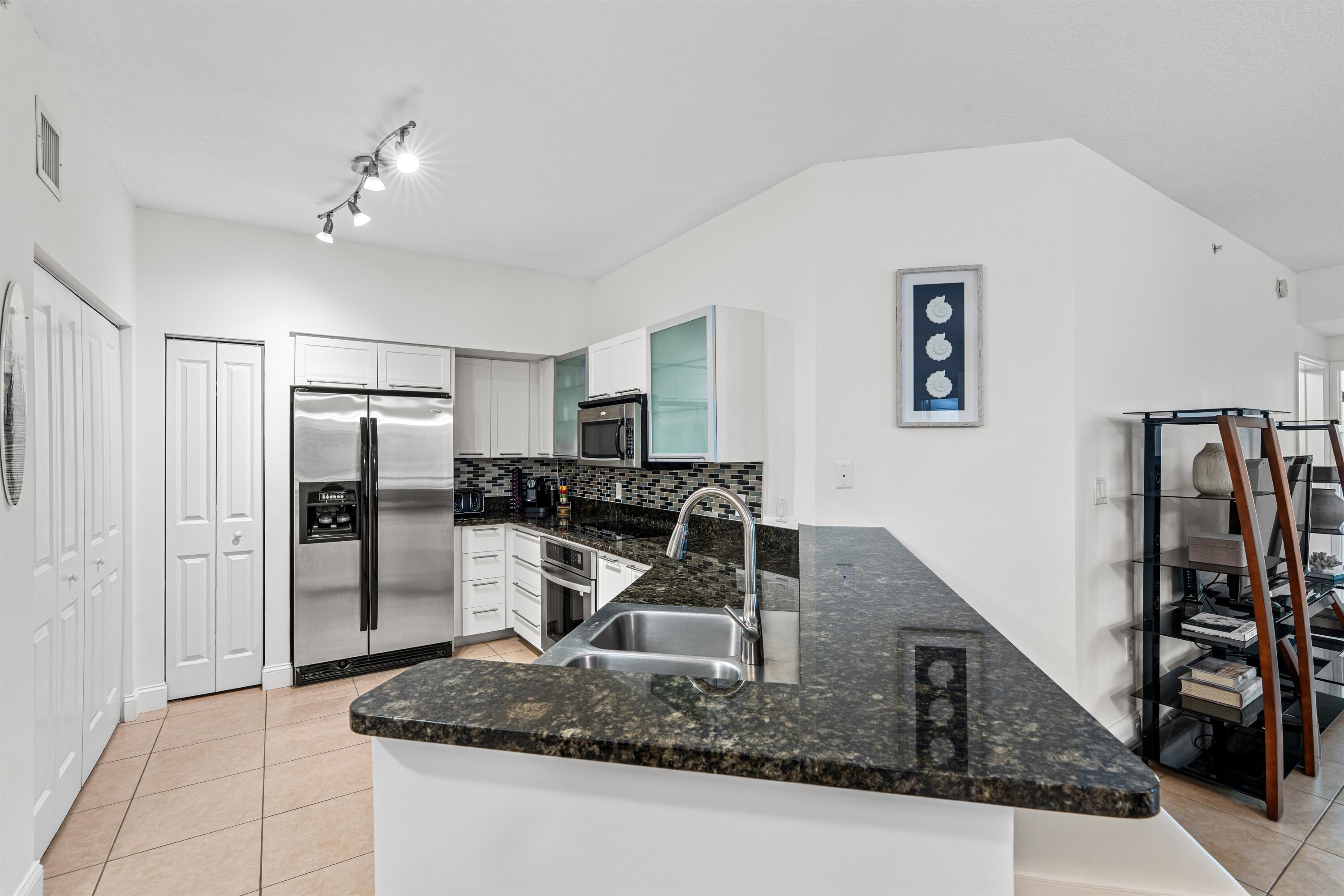 Kitchen featuring a stone-surface breakfast bar, mosaic tile backsplash, stainless steel appliances, white cabinetry with glass-front accents, and tile flooring