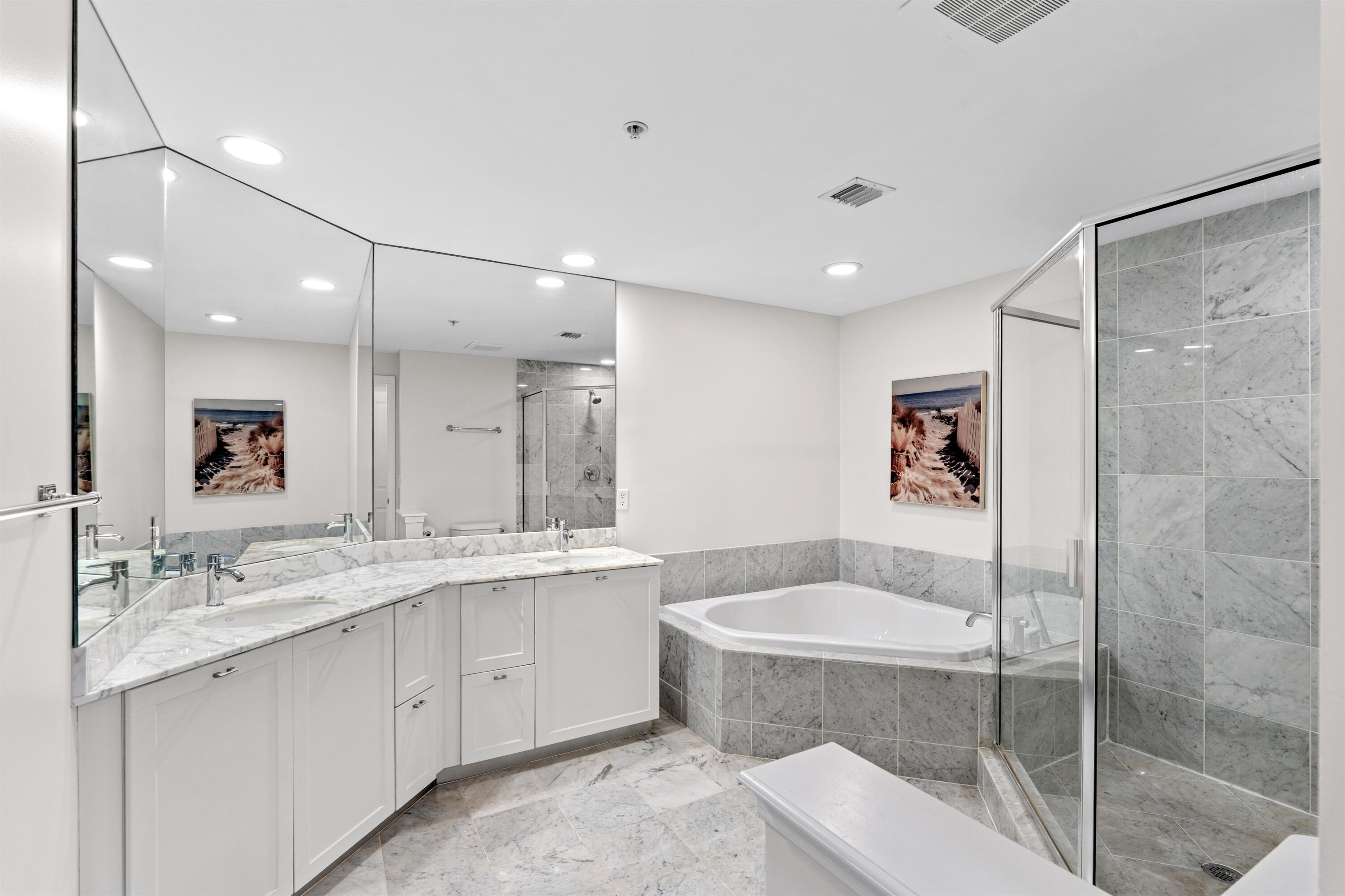 White cabinetry with stone countertops, expansive wall-to-wall mirror, corner soaking tub, stone-tiled walk-in shower with glass enclosure, and stone flooring