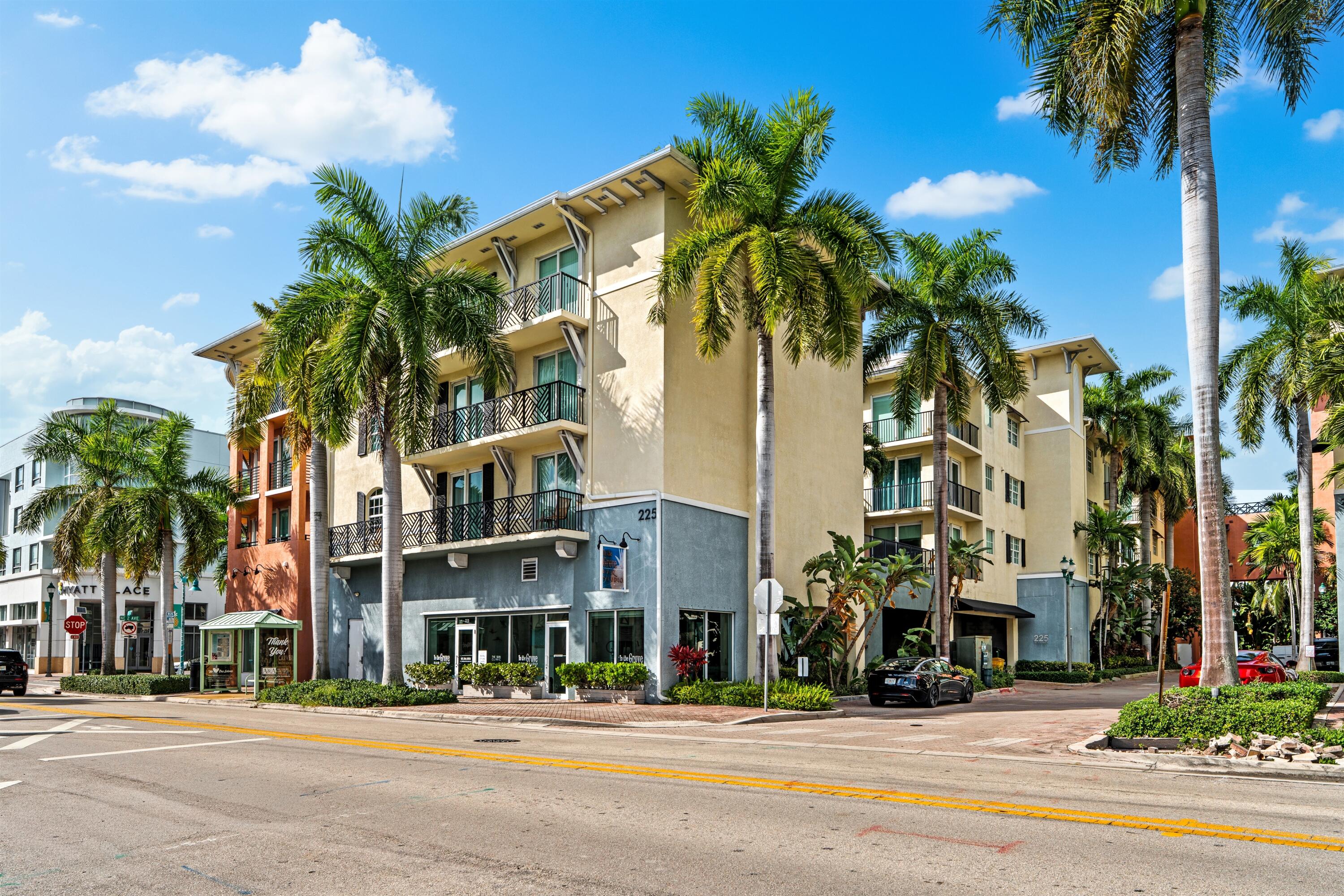 Multi-story stucco building exterior featuring metal balcony railings, prominent palm tree landscaping, and floor-to-ceiling ground-level windows