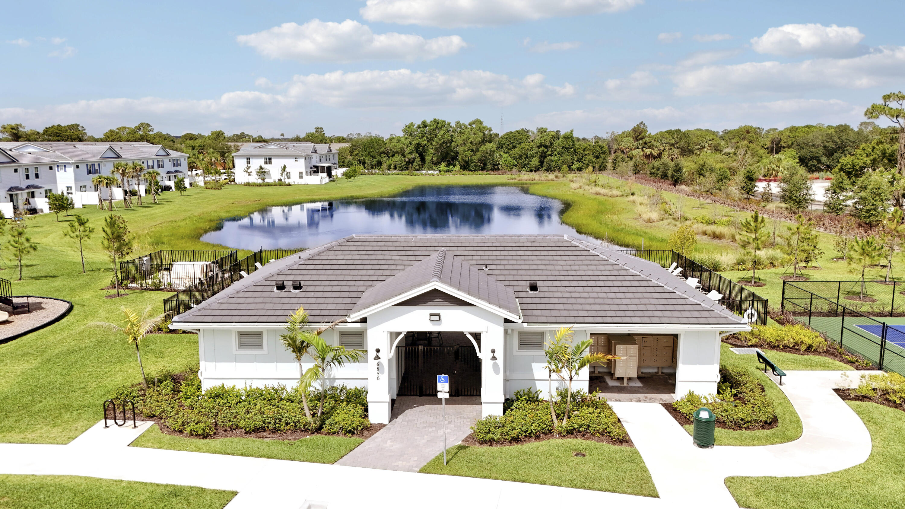 Community clubhouse featuring a dark tile roof, exterior lighting fixtures, and a paved walkway, situated on grounds with a retention pond and sport court.