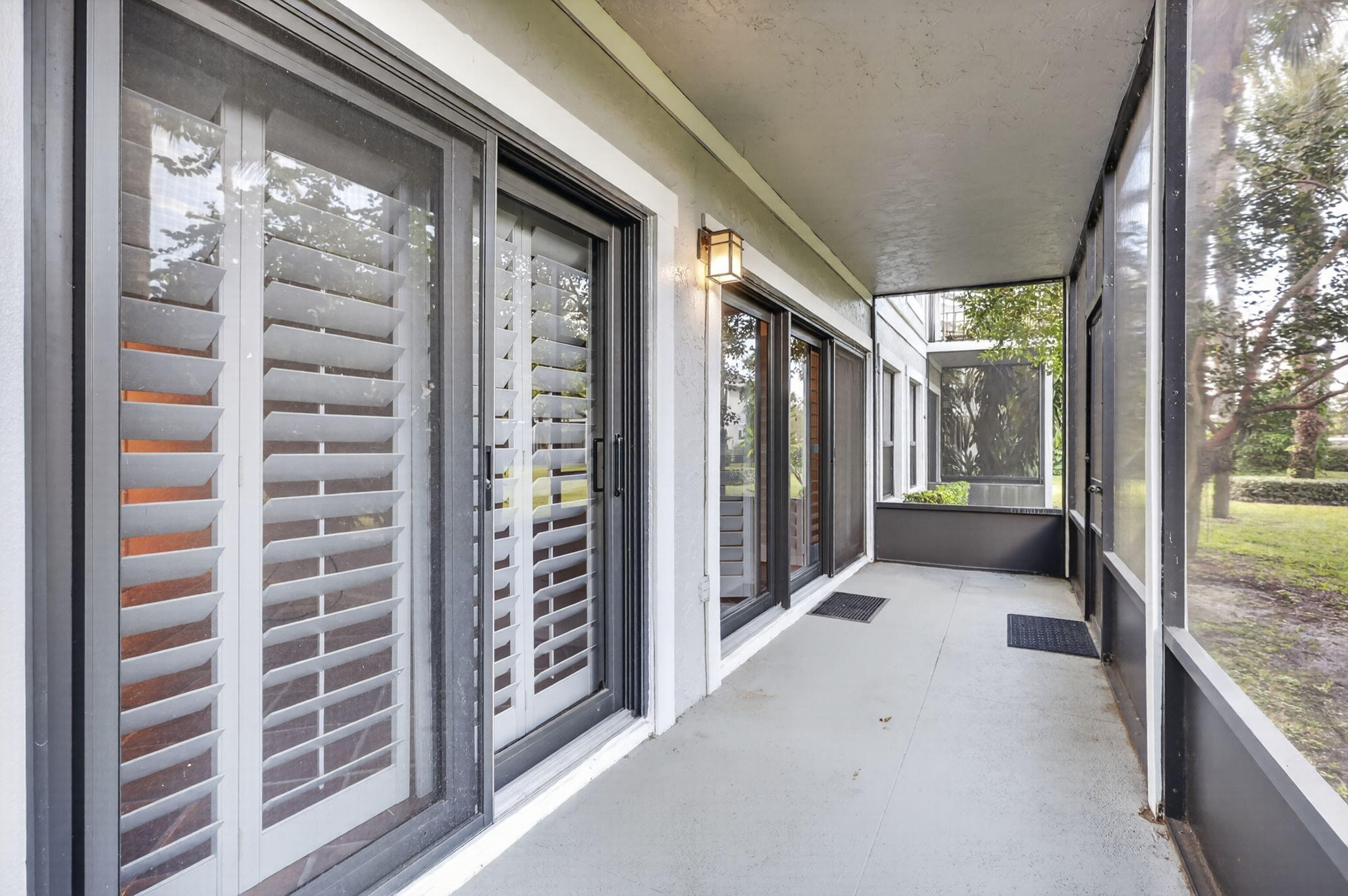 Screened-in porch featuring concrete flooring, dark metal framing, sliding glass doors with white plantation shutters, and a wall-mounted exterior light fixture