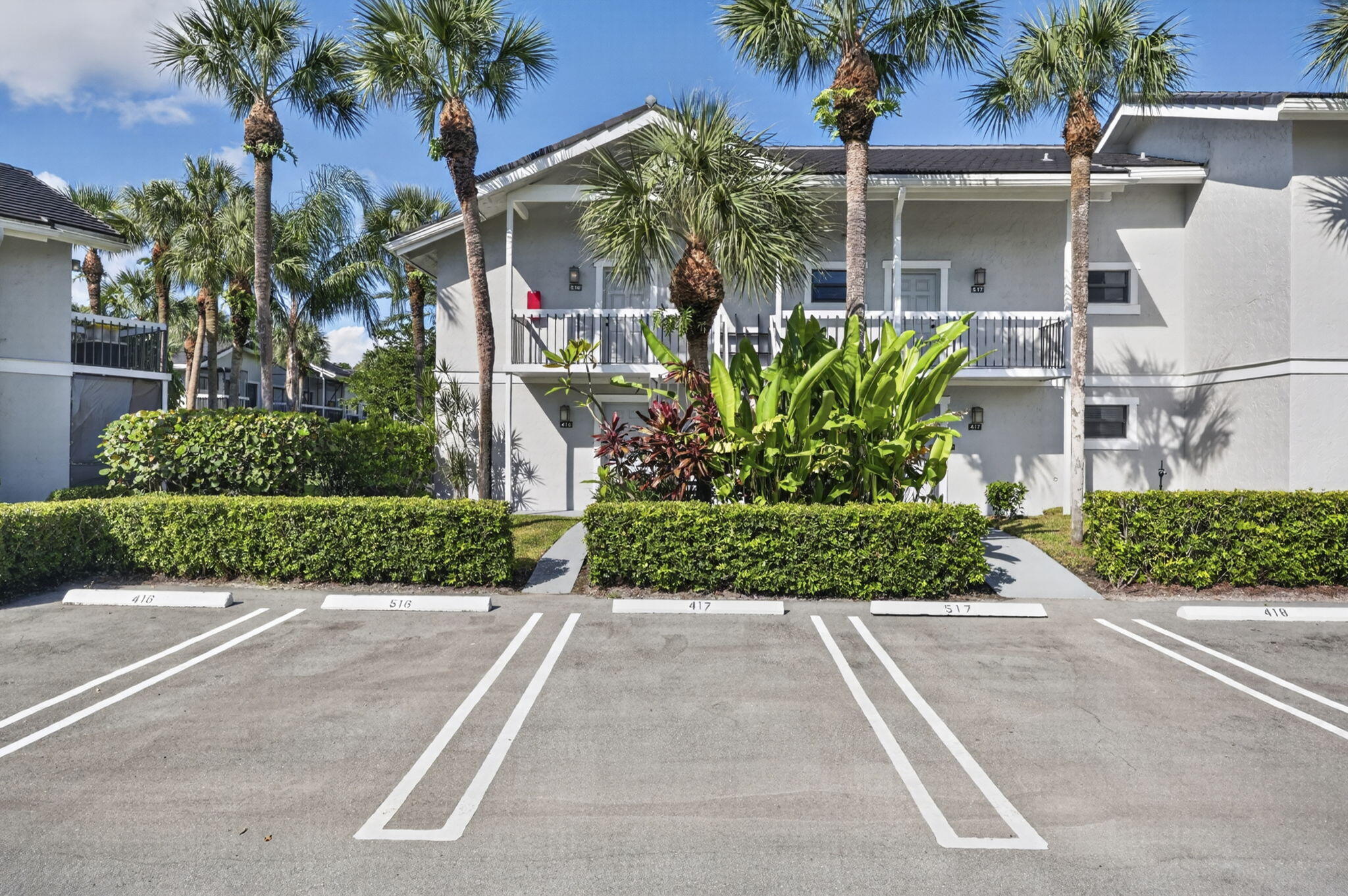 Two-story exterior featuring a stucco facade, balcony with white railings, mature palm trees, and manicured shrubbery