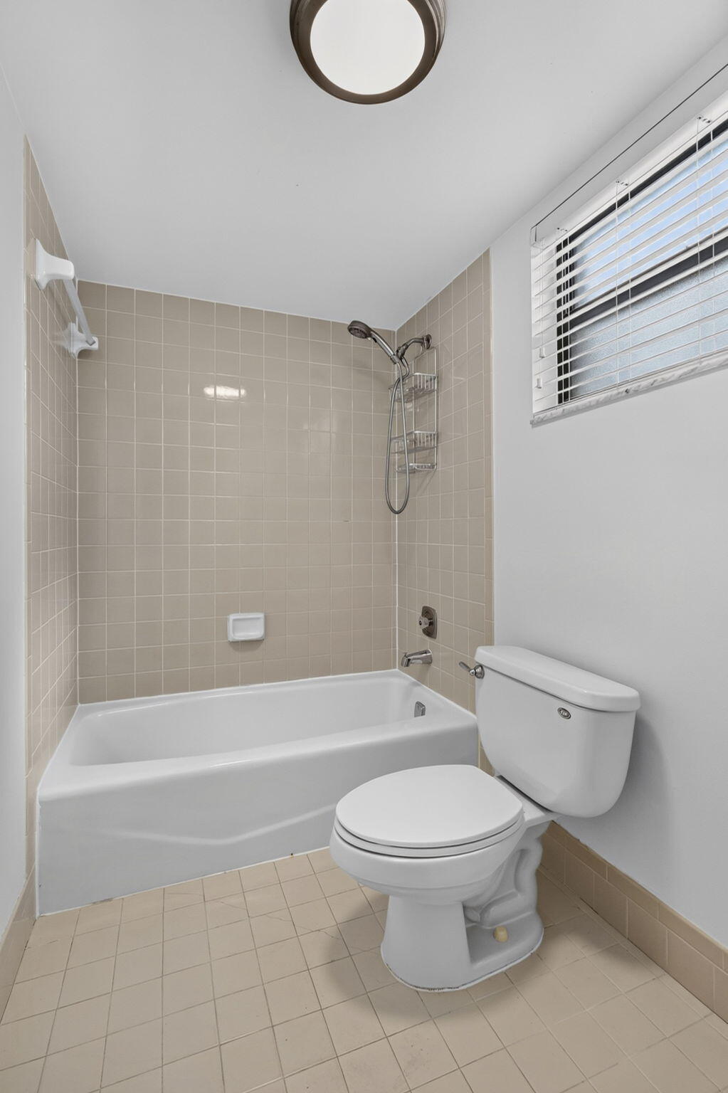 Bathroom featuring a built-in bathtub with a tile surround, window with horizontal blinds, flush-mount lighting, and neutral floor tiling