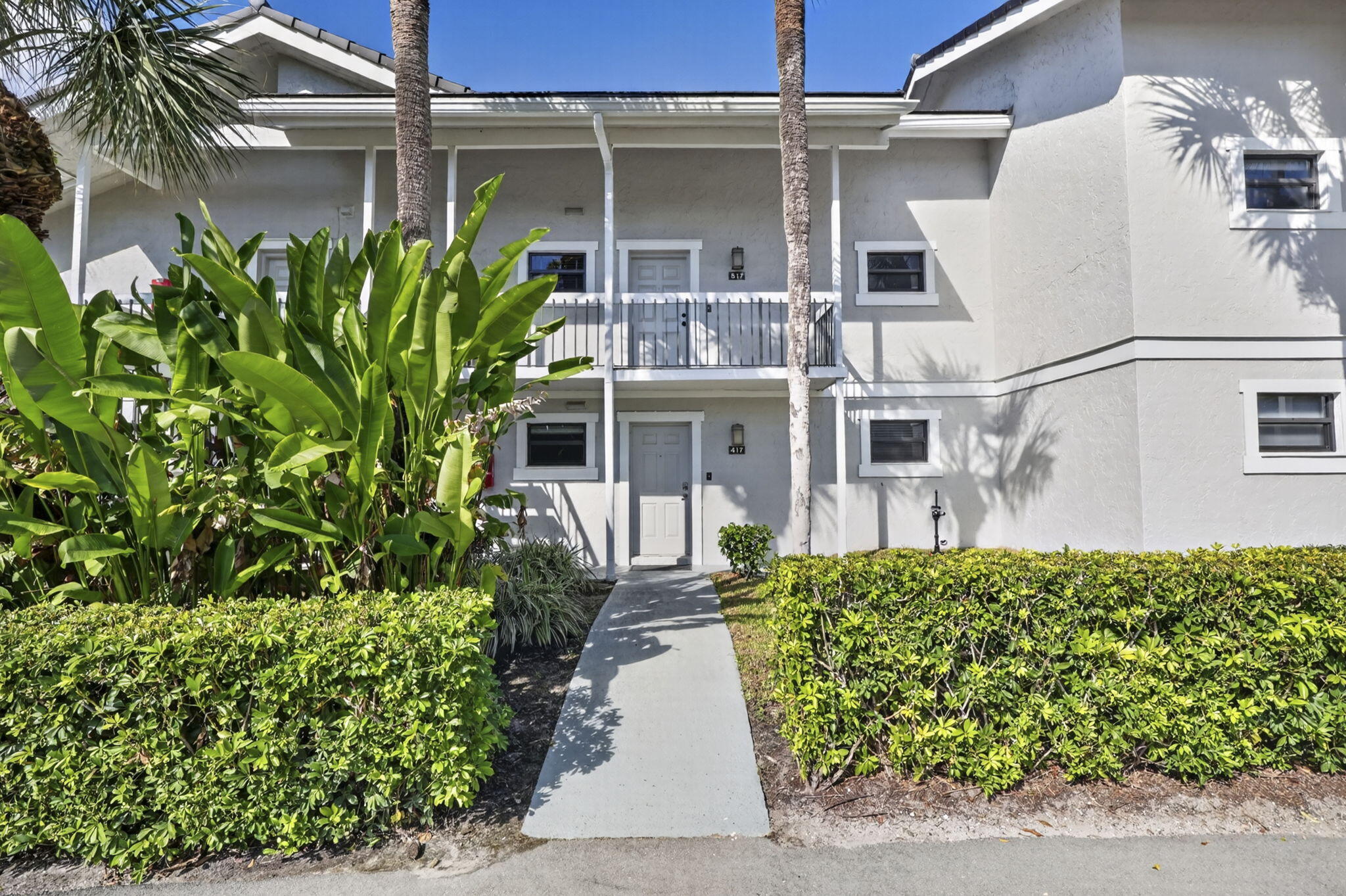 Two-story exterior featuring light-toned stucco siding, a concrete walkway, a second-level balcony with metal railing, and mature ornamental shrubbery Two-story exterior featuring light-toned stucco siding, a concrete walkway, a second-level balcony with metal railing, and mature ornamental shrubbery