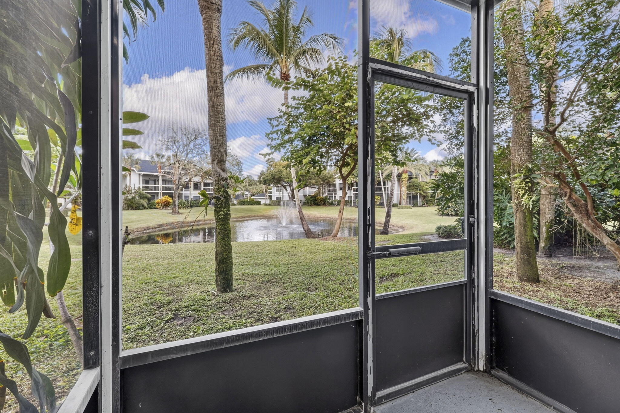 Screened-in enclosure featuring a metal-framed door, concrete flooring, exterior foliage, and a pond with a fountain