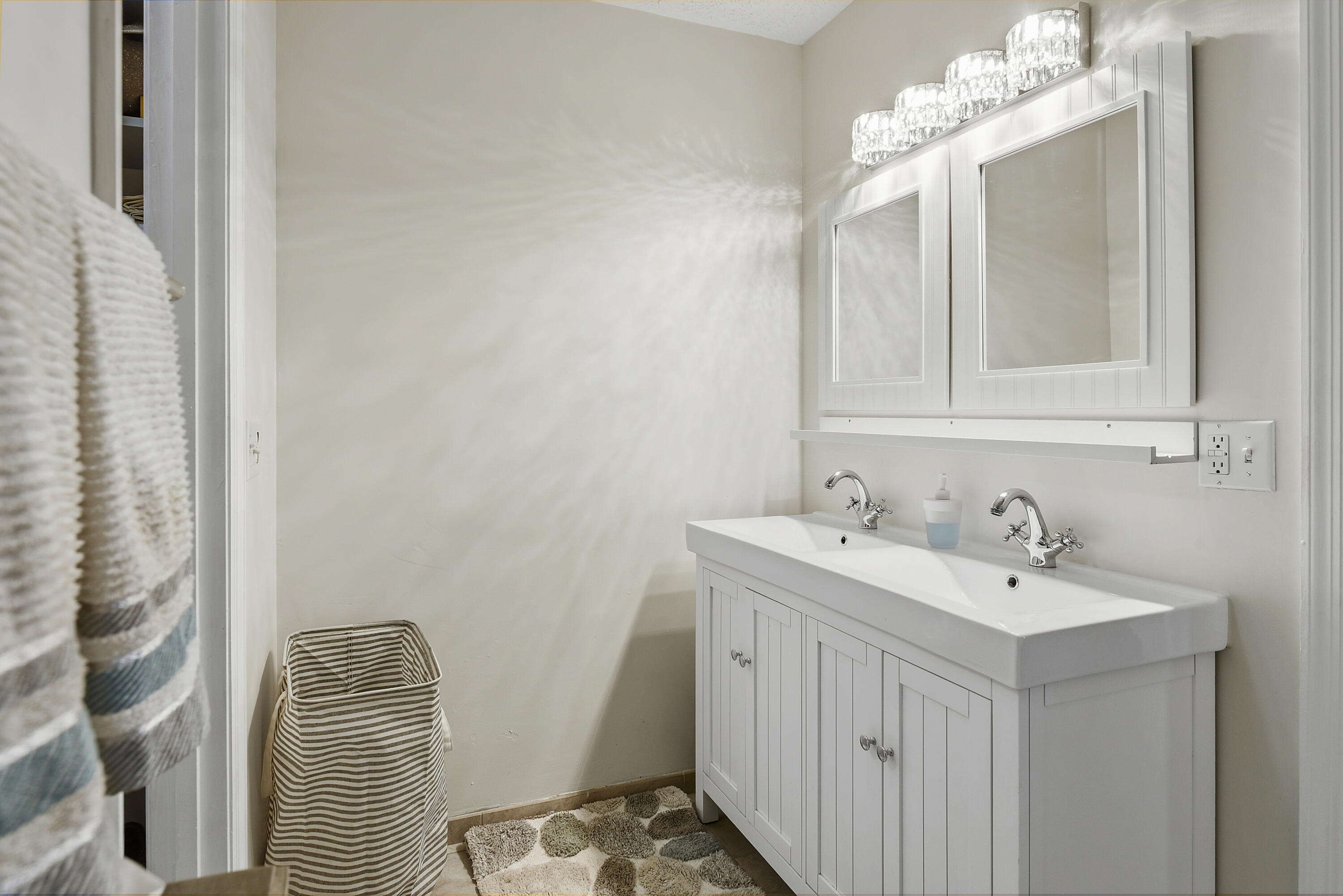 Bright primary bathroom featuring a double-sink vanity, decorative mirrors, updated lighting, and convenient access to two separate walk-in closets.