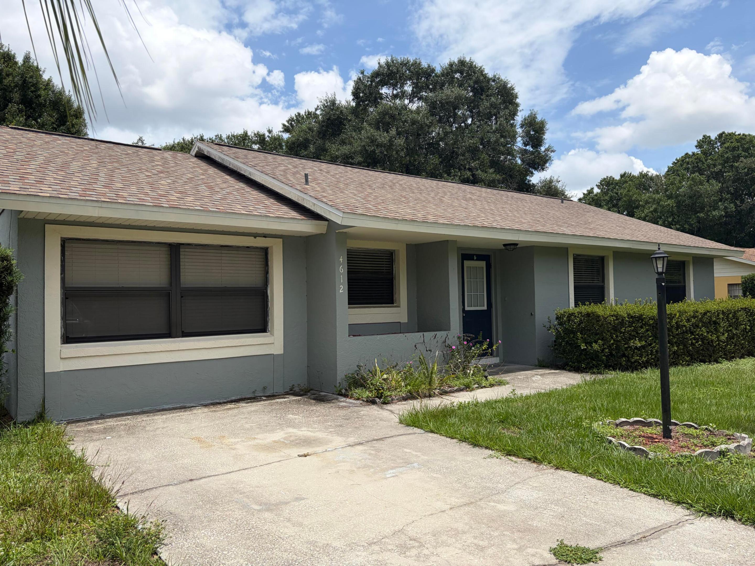 Single-story exterior featuring a neutral-toned facade, shingled roof, concrete driveway, and attached exterior lighting fixture Single-story exterior featuring a neutral-toned facade, shingled roof, concrete driveway, and attached exterior lighting fixture