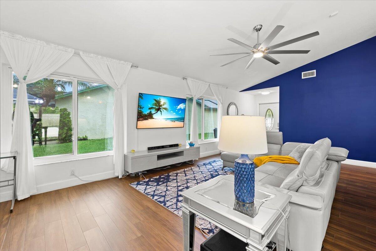 Spacious room featuring wood-finish flooring, a vaulted ceiling with a multi-blade ceiling fan, large windows, and an accent wall
