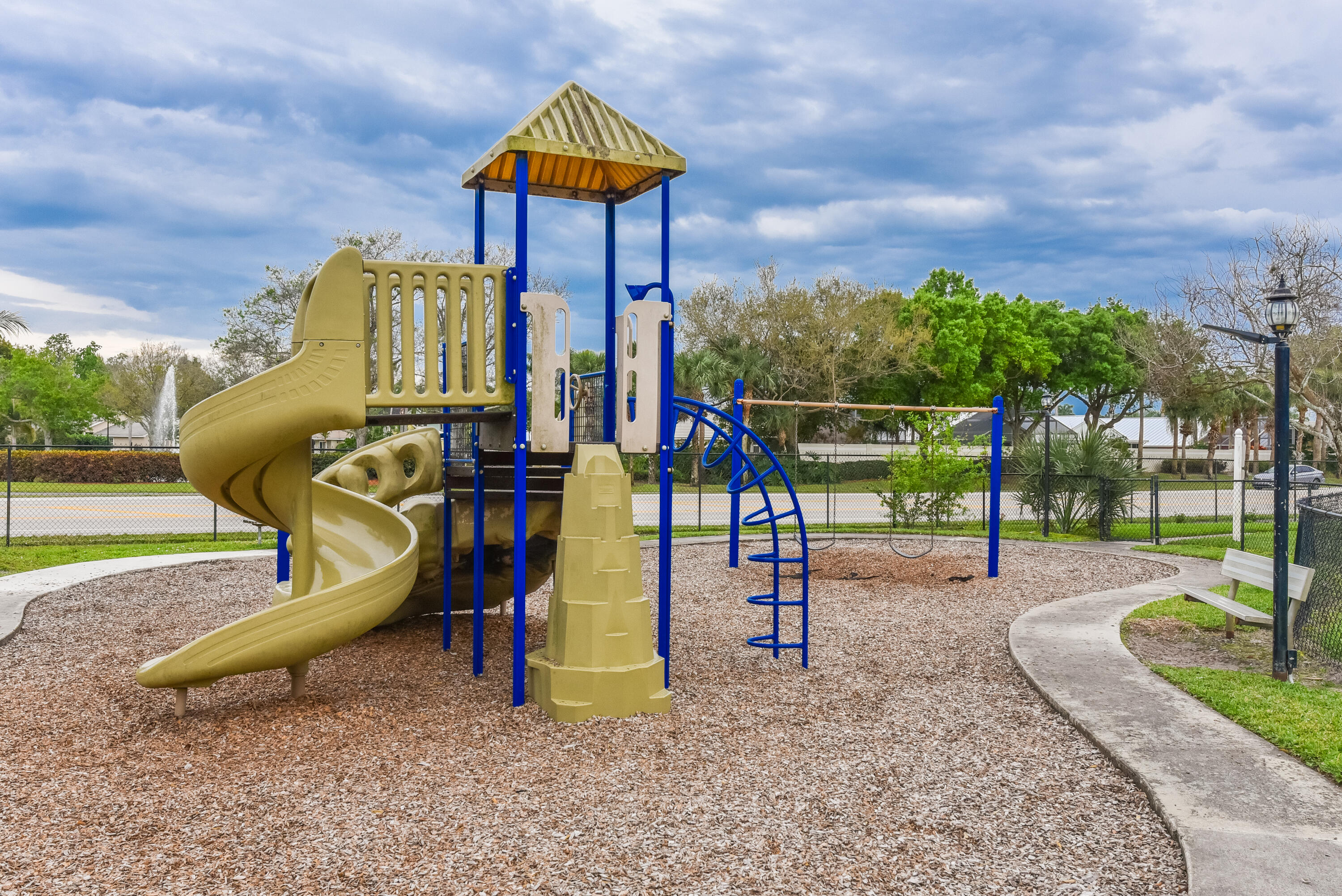 Outdoor recreation area featuring a multi-level climbing structure with a spiral slide, swing set, wood chip ground cover, and concrete perimeter walkway