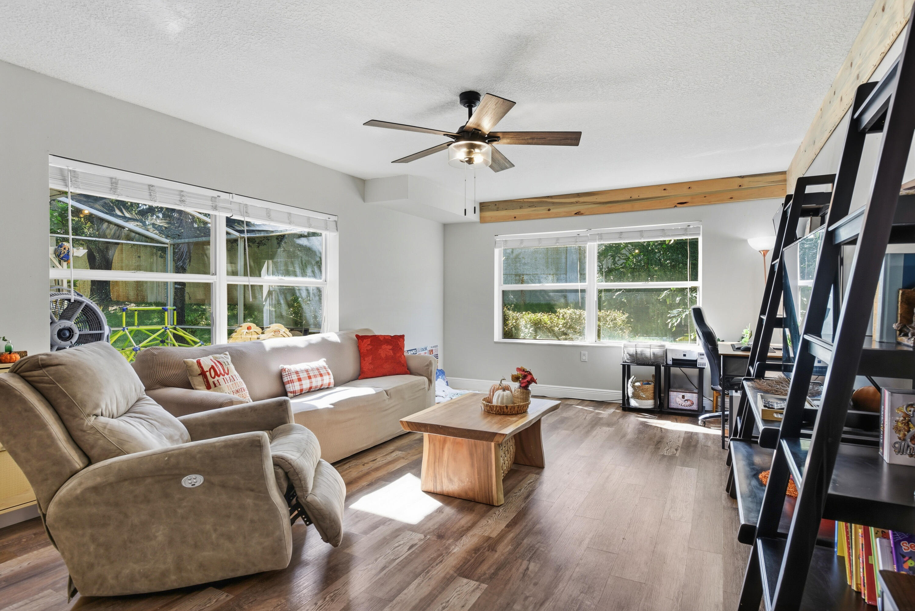 Living area featuring wood-finish flooring, exposed natural wood ceiling beams, a ceiling fan with integrated lighting, and multiple windows