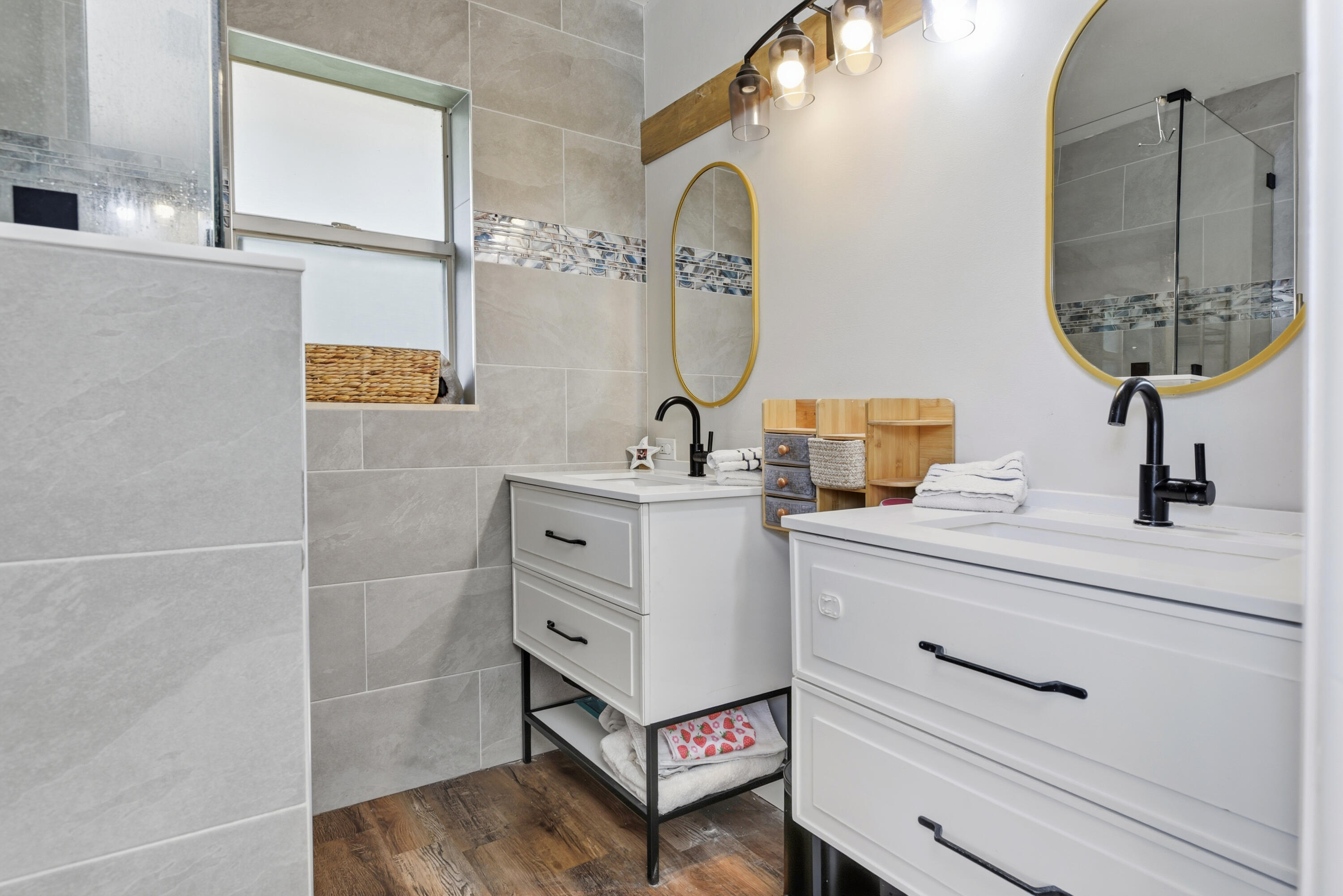 Dual vanity with white cabinetry, matte black hardware, integrated rectangular sinks, and wood-finish flooring, complemented by neutral oversized wall tile and arched mirrors with gold-tone frames