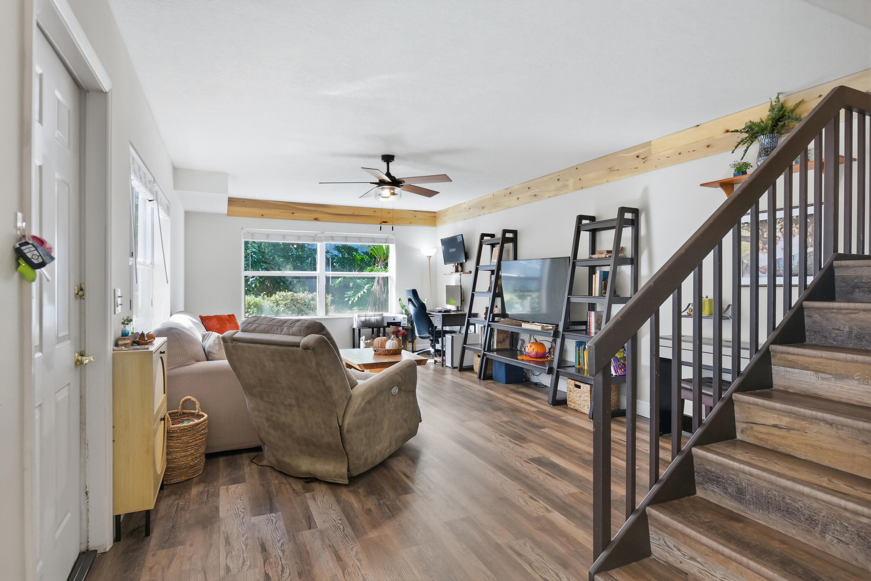 Living area featuring wood-finish flooring, exposed natural wood ceiling beams, a ceiling fan with integrated lighting, and a staircase with dark wood handrails and balusters