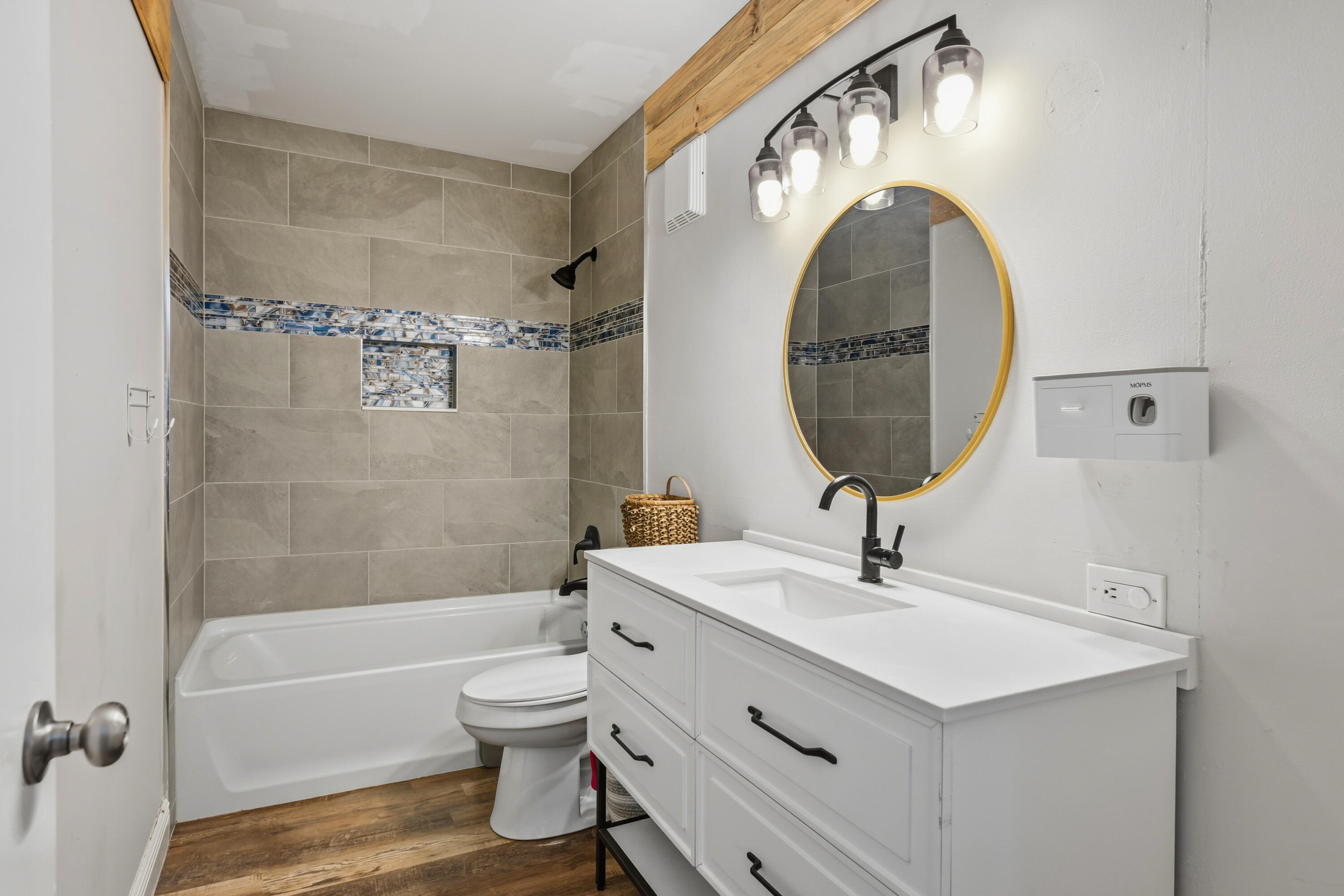 Bathroom featuring wood-finish flooring, white vanity with matte black hardware, round mirror with metallic trim, and tub-shower combination with decorative tile inlay