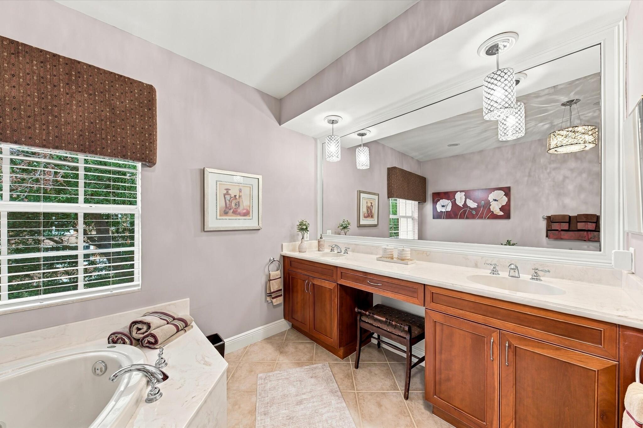 Dual-sink vanity with wood-finish cabinetry, solid surface countertop, soaking tub with tile surround, full-width mirror, and hanging pendant lighting
