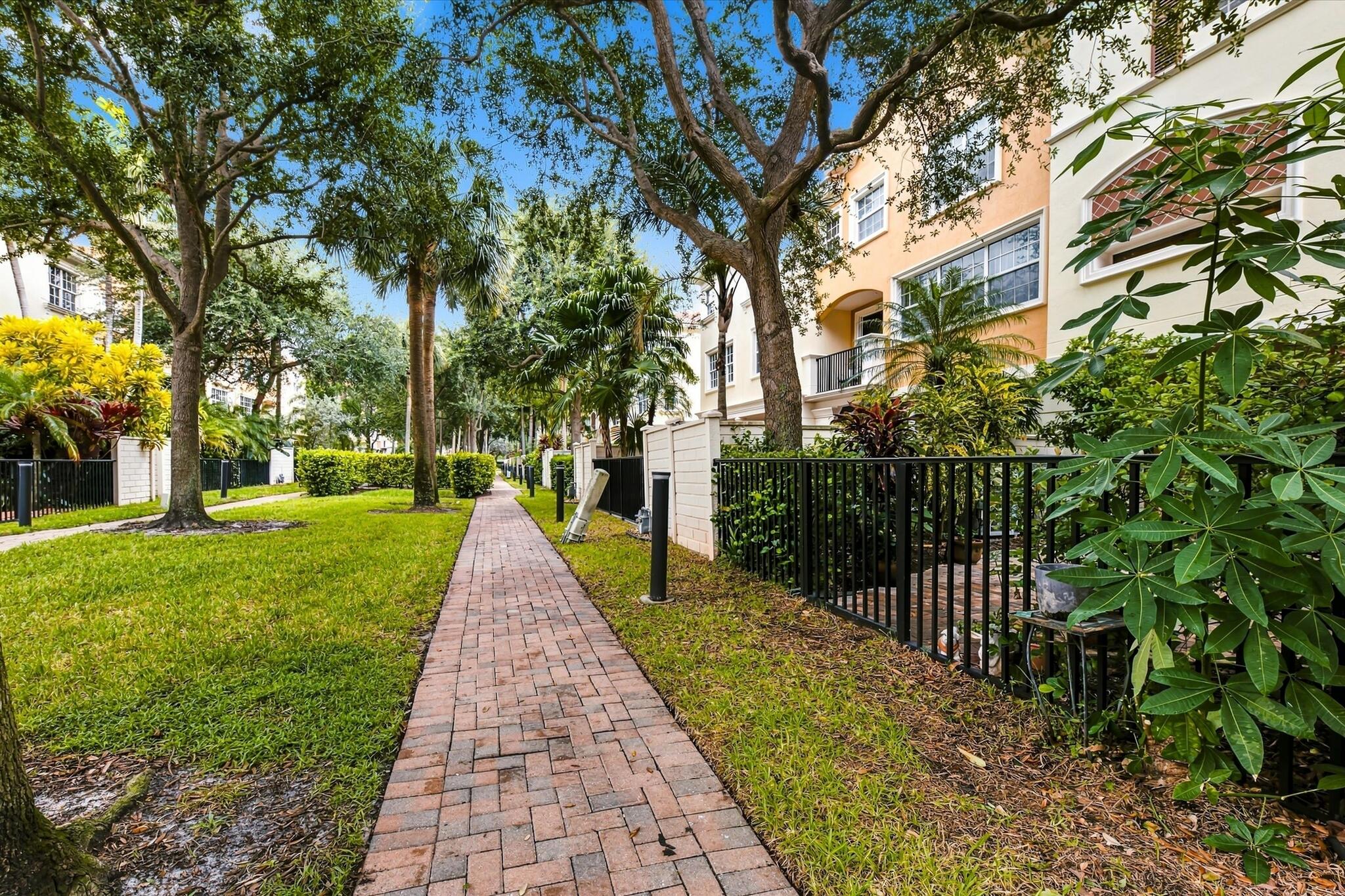 Brick paver walkway lined with mature trees, established landscaping, and black metal fencing