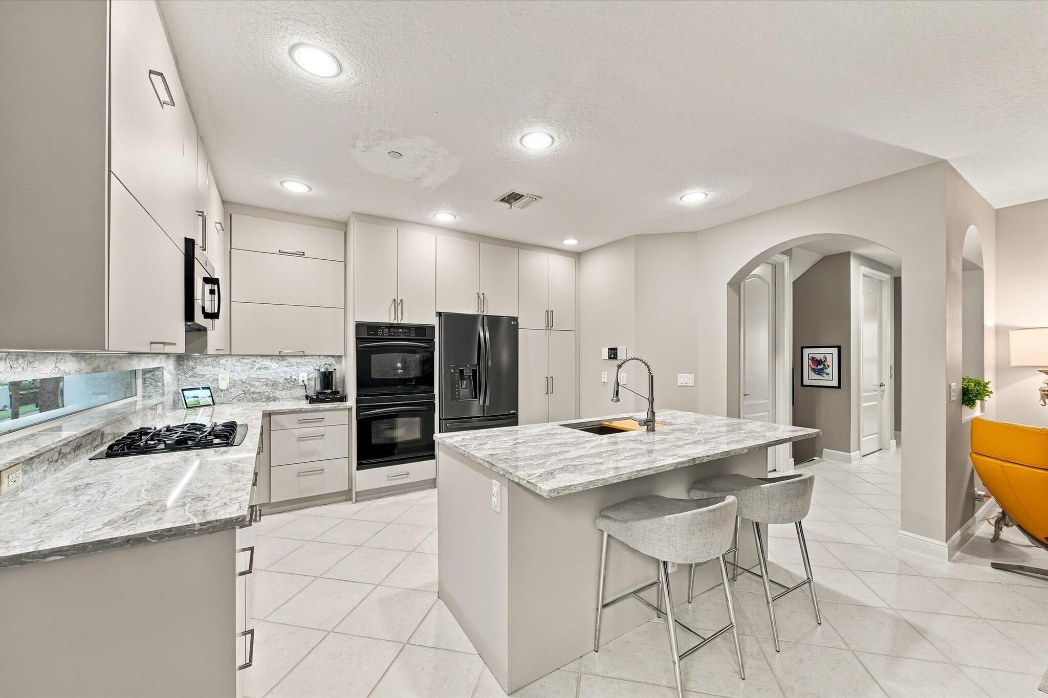 Kitchen featuring light-toned cabinetry, stone countertops, a central island with an integrated sink, built-in double ovens, and recessed lighting