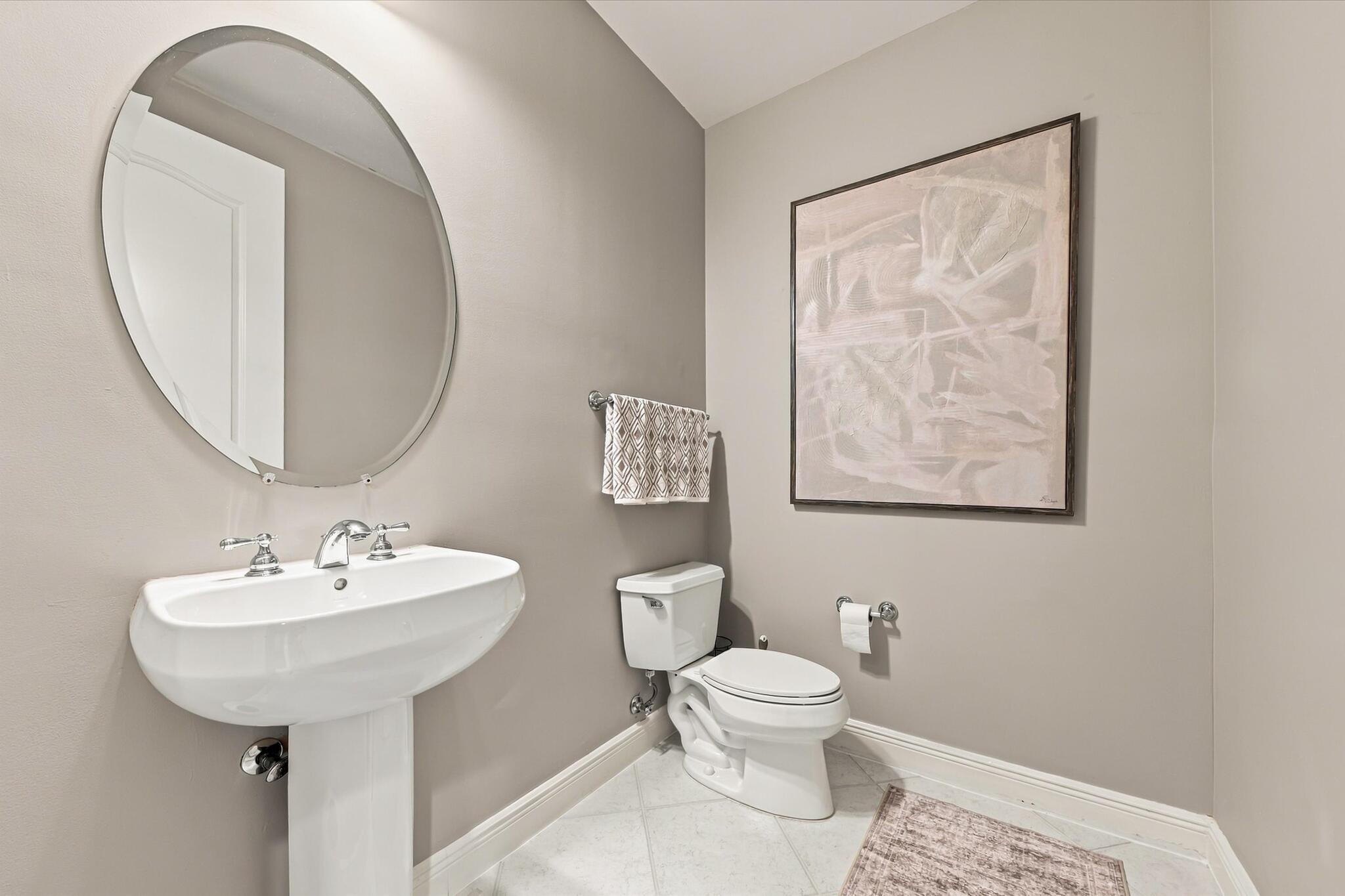 Oval mirror with a metallic frame, white porcelain pedestal sink, polished chrome widespread faucet, and ceramic tile flooring