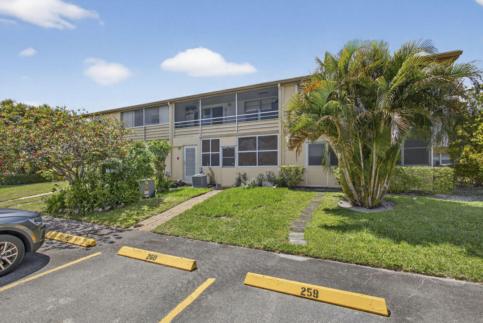 Two-story multi-family exterior featuring light-toned siding, second-level screen-enclosed balconies, ground-level screen-enclosed patio, and established tropical landscaping