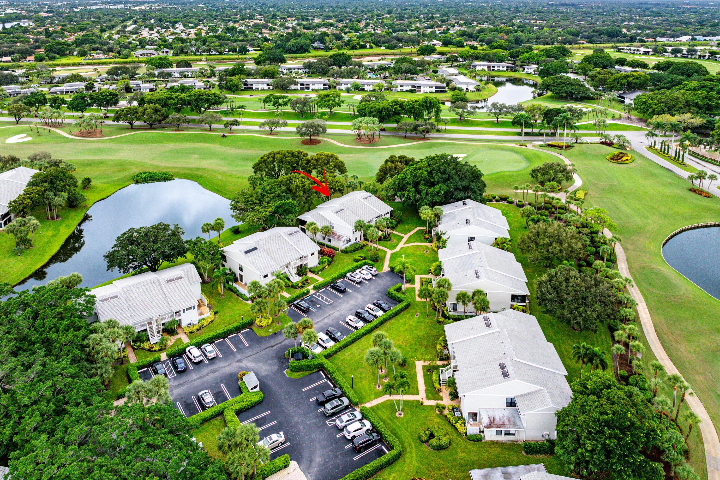 Aerial perspective of a multi-unit residential complex featuring light-toned exteriors, pitched roofs, paved surface parking, and manicured grounds integrated with water features and golf course fairways
