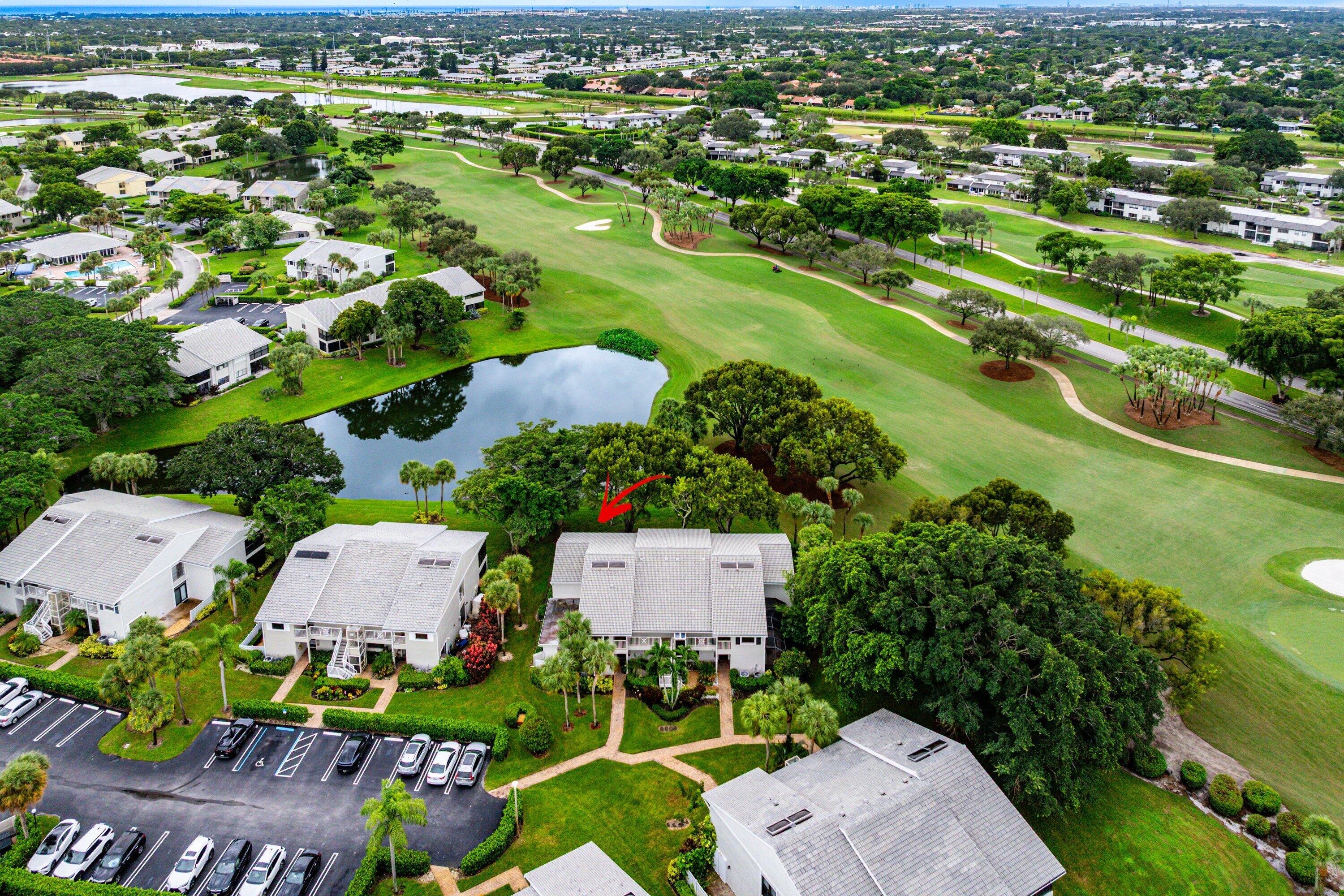 Aerial perspective featuring multi-story residential buildings with light-toned shingle roofing, positioned along a water feature and bordering an expansive manicured green space