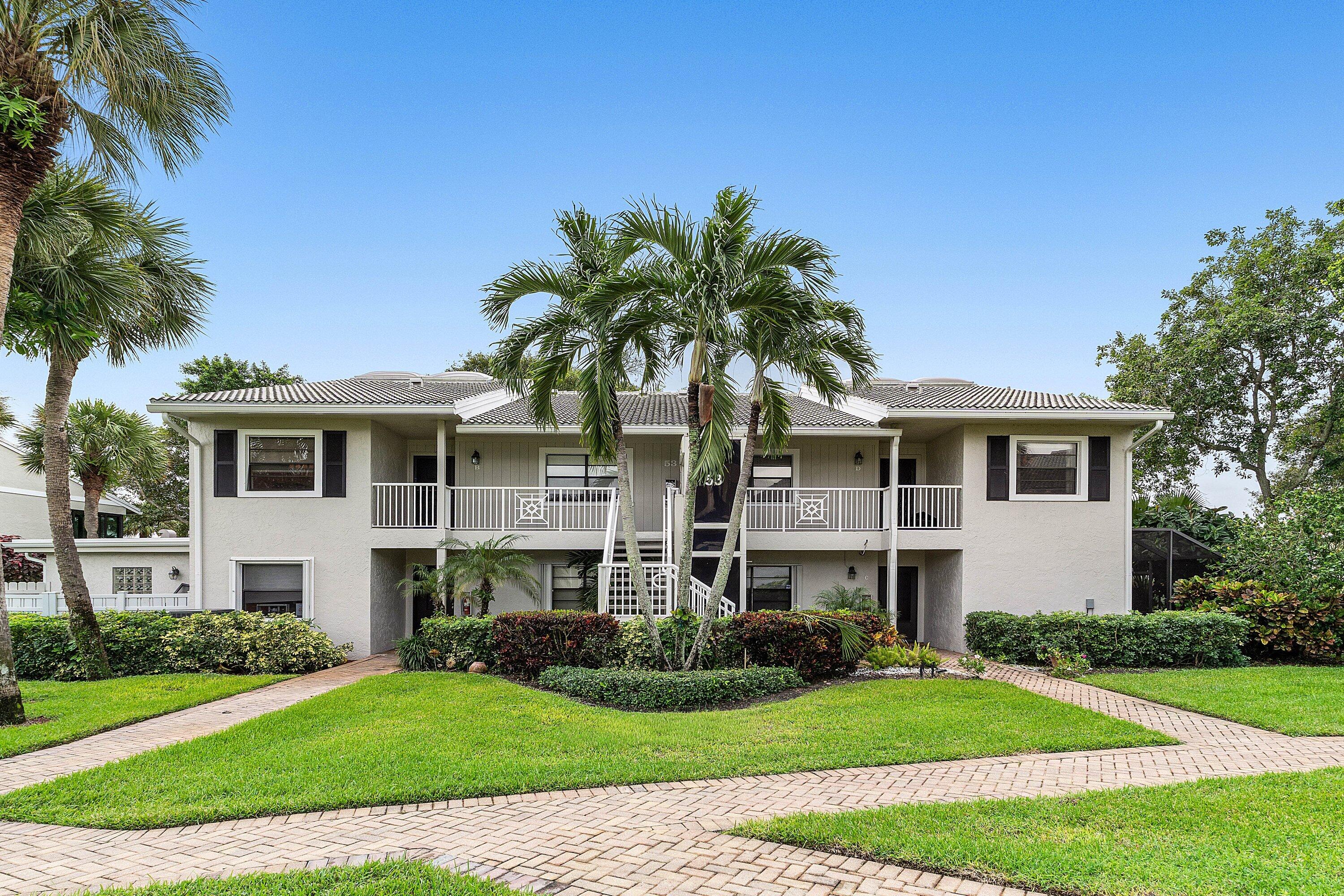 Two-story exterior featuring light-toned stucco siding, a tiled roof, upper-level balconies with white railings, and paver walkways surrounding a manicured lawn