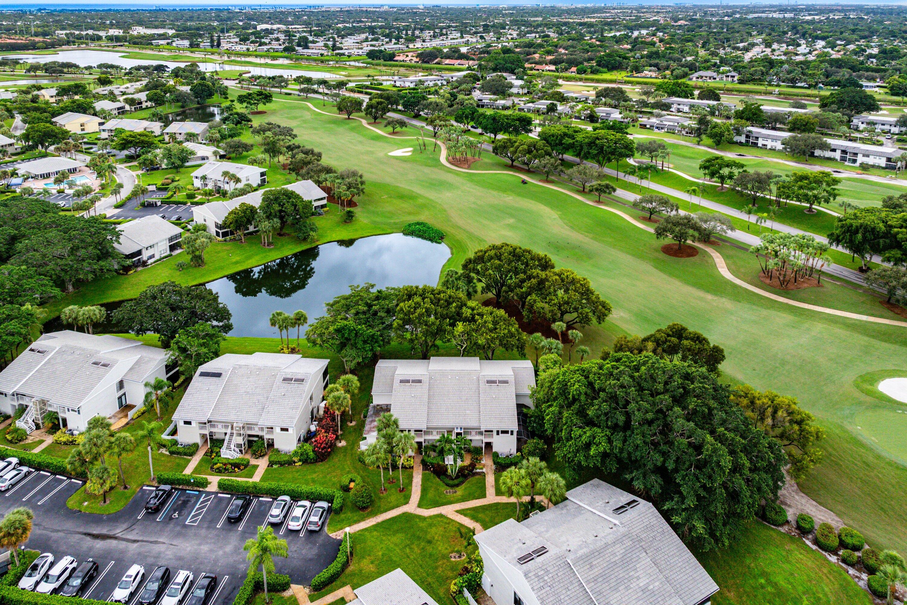 Aerial perspective of multi-family residences featuring light-colored shingled roofs, landscaped grounds with mature trees, a central pond, and proximity to golf course fairways