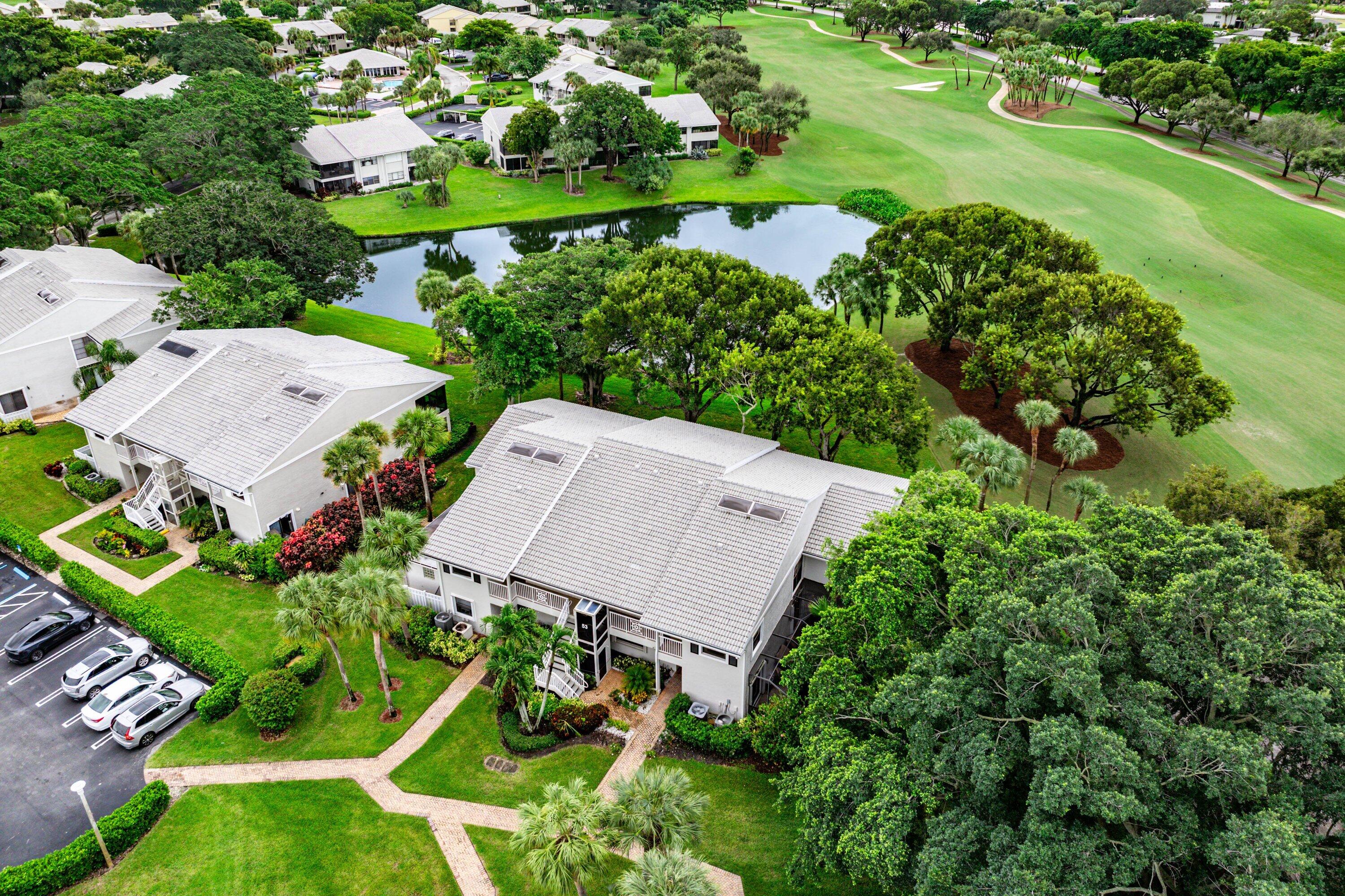 Aerial perspective of multi-unit residential buildings featuring light-toned roof shingles, skylights, paver walkways, and mature landscaping situated along a pond and golf course fairways
