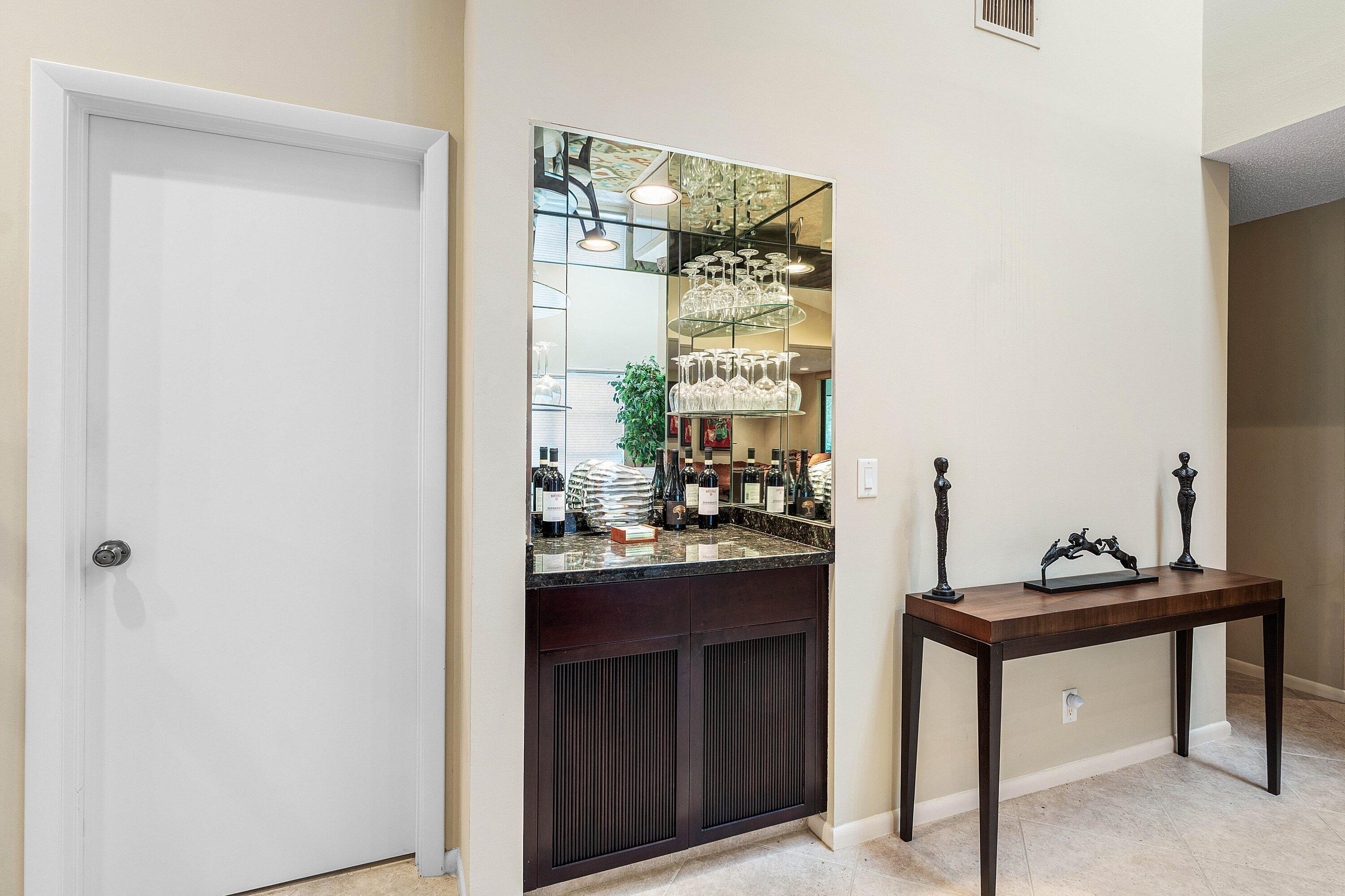 Built-in wet bar featuring a stone countertop, dark wood cabinetry with slatted panel doors, integrated mirrored backsplash, and overhead glass shelving