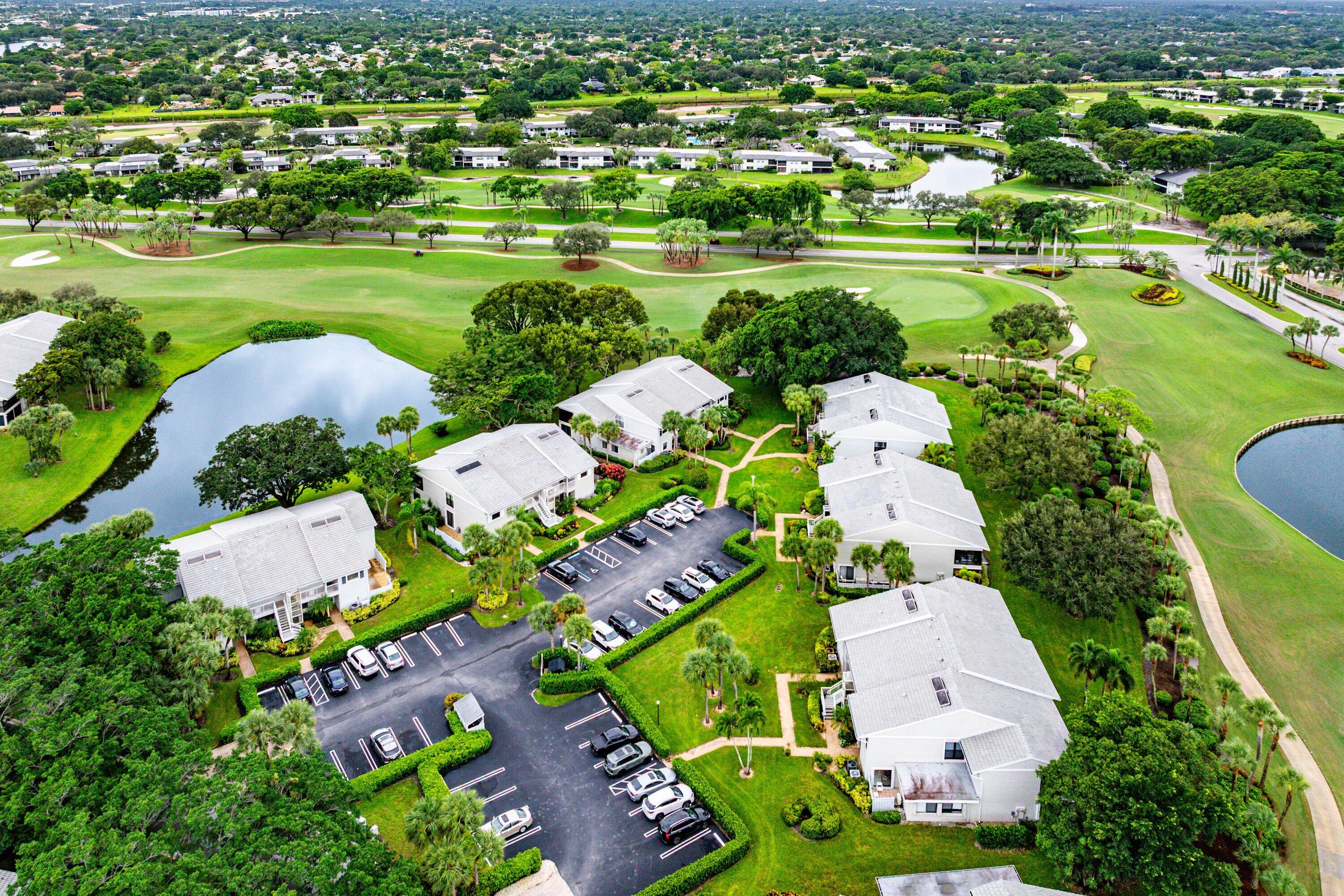 Aerial perspective of residential structures featuring light-toned pitched roofs, paved surface parking, manicured grounds, mature trees, and direct proximity to a golf course and water features