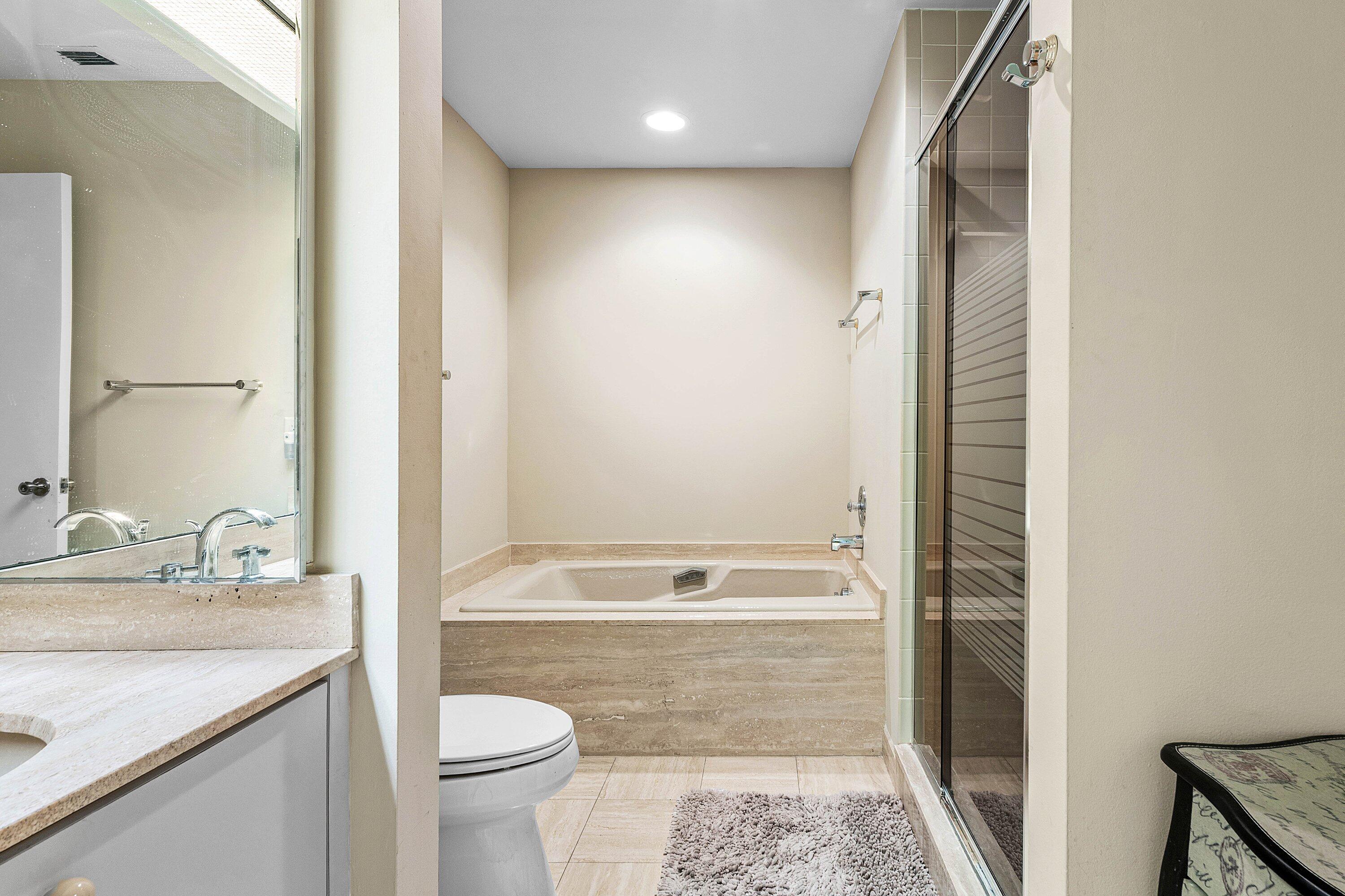 Bathroom featuring a tiled soaking tub with a stone-surround deck, separate glass-enclosed shower, vanity with stone countertop, and wall-to-wall mirror