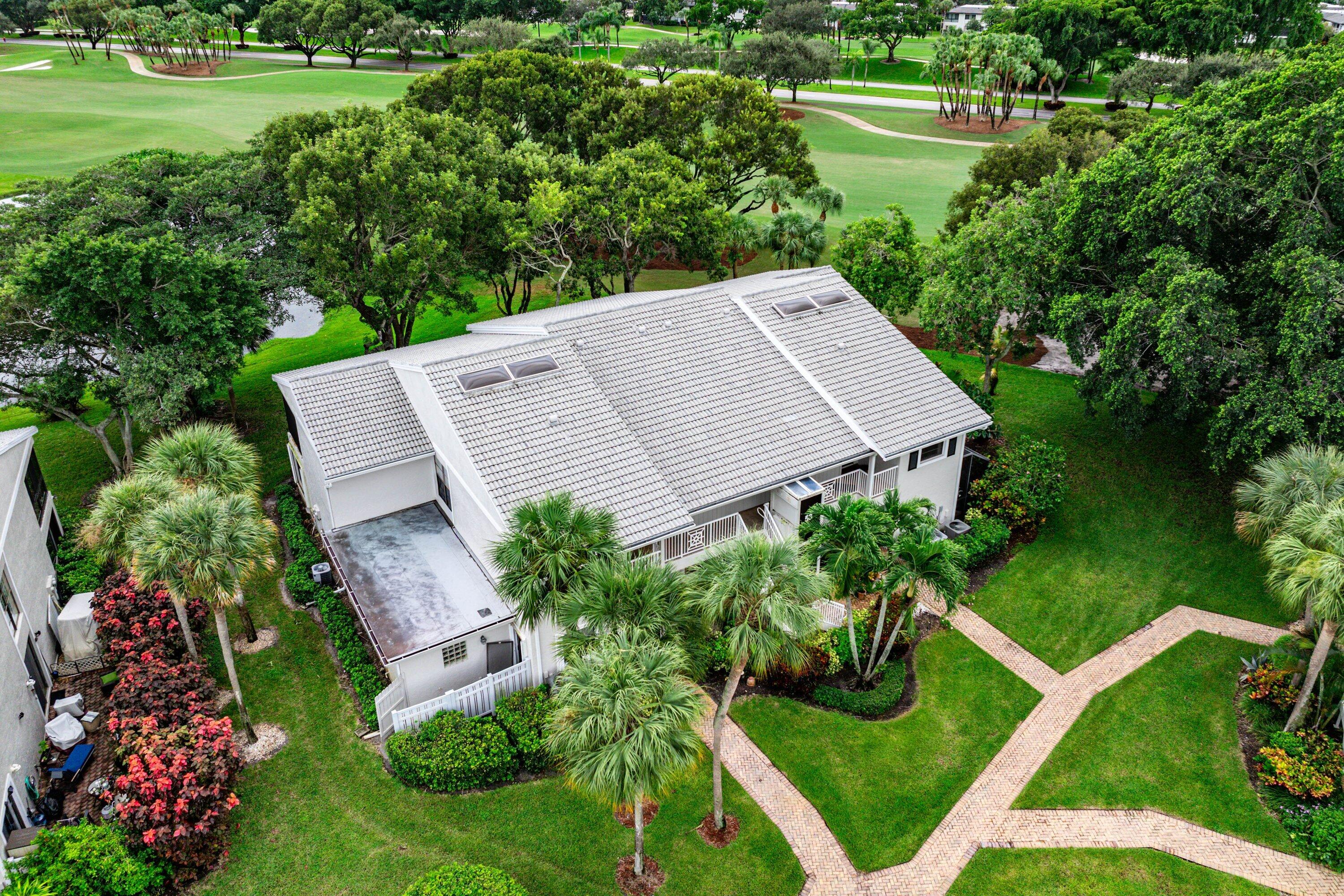 Exterior aerial featuring a multi-pitched roof with integrated skylights, a light-toned facade, mature palm trees, and manicured landscaping with paver walkways