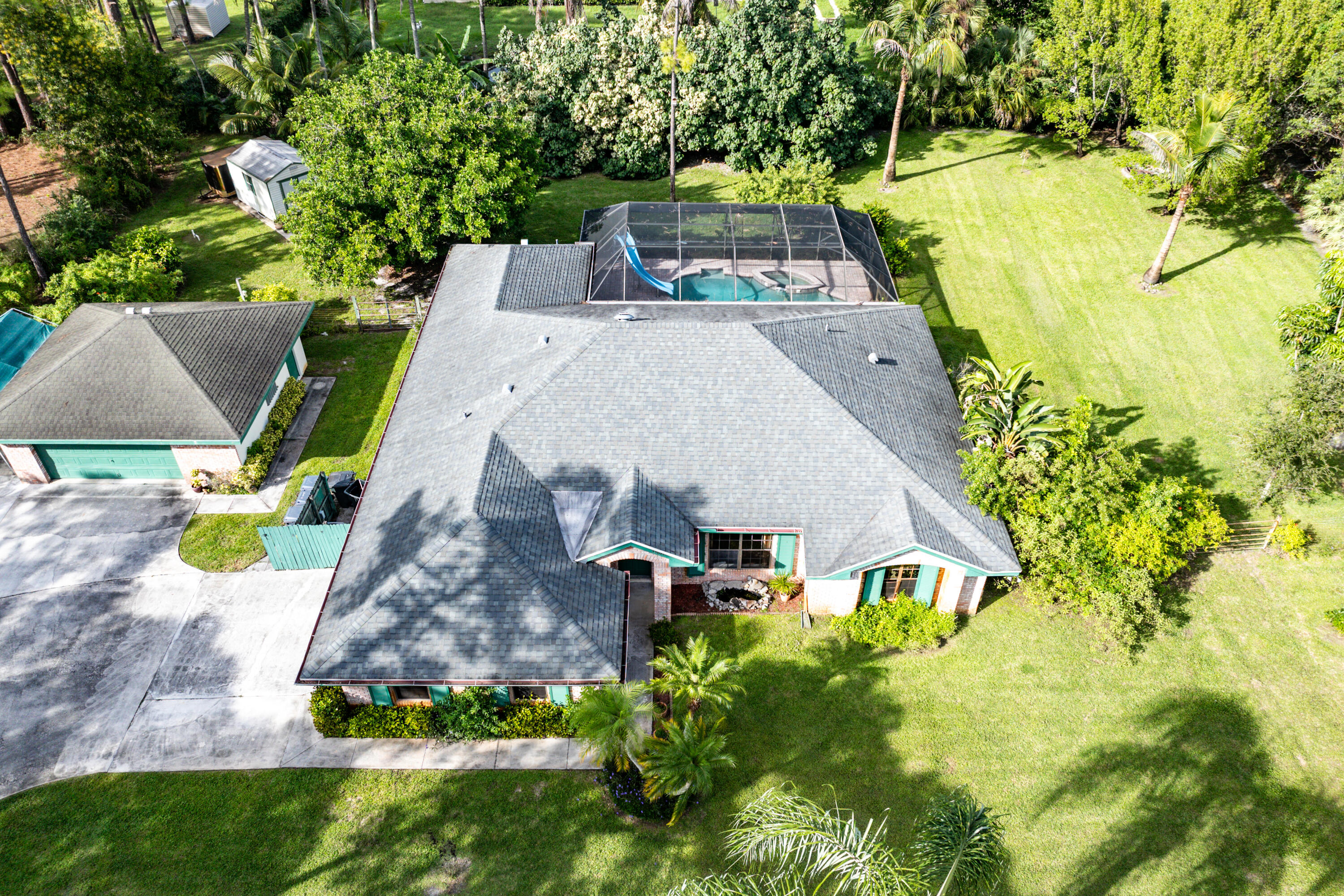 Aerial perspective featuring a multi-gabled shingle roof, expansive manicured grounds, a screened-in pool enclosure with an integrated water slide, a detached garage, and an extensive concrete driveway