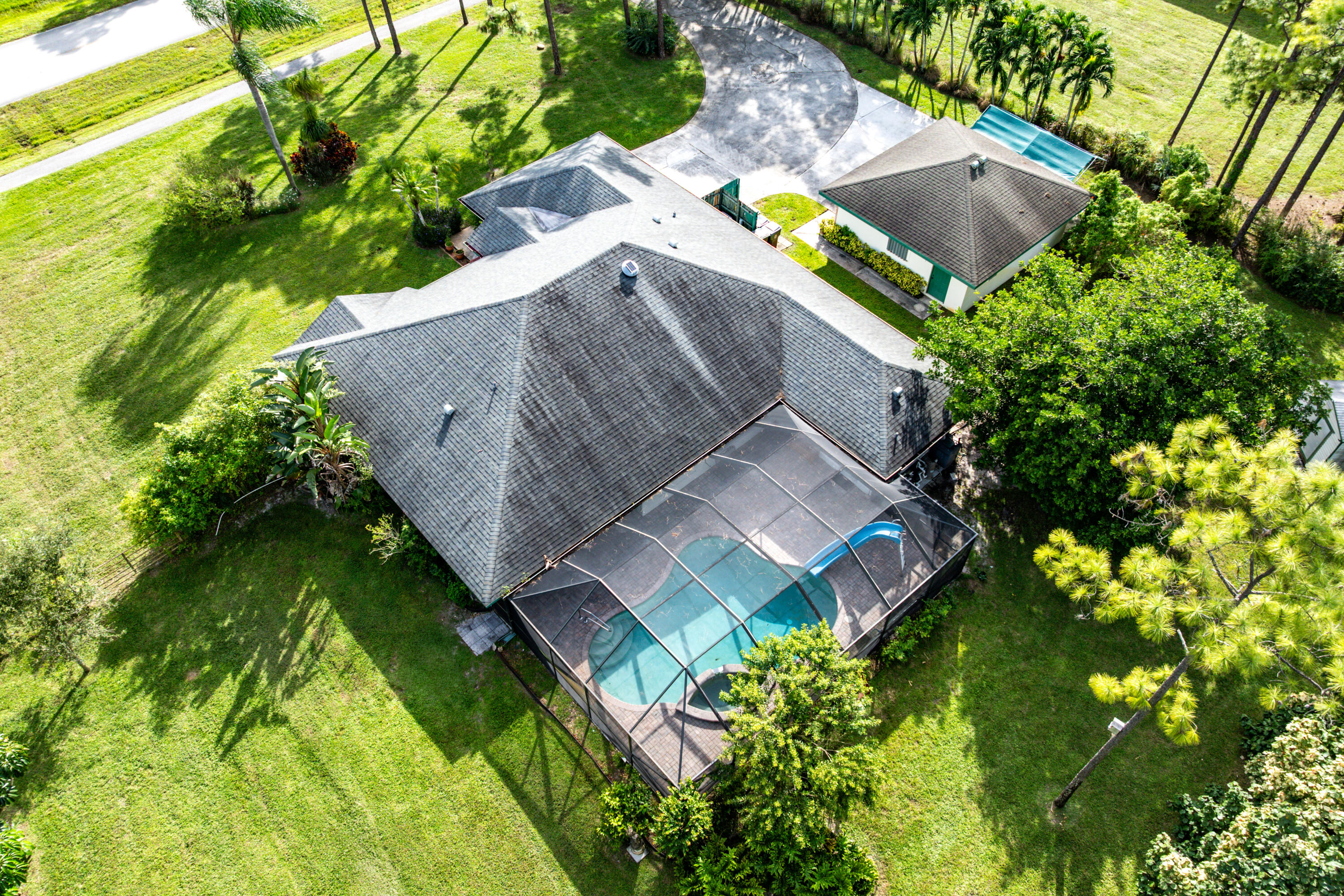 Aerial perspective of a sprawling property featuring a screened-in pool enclosure, a detached structure, a paved driveway, and expansive grounds with mature trees