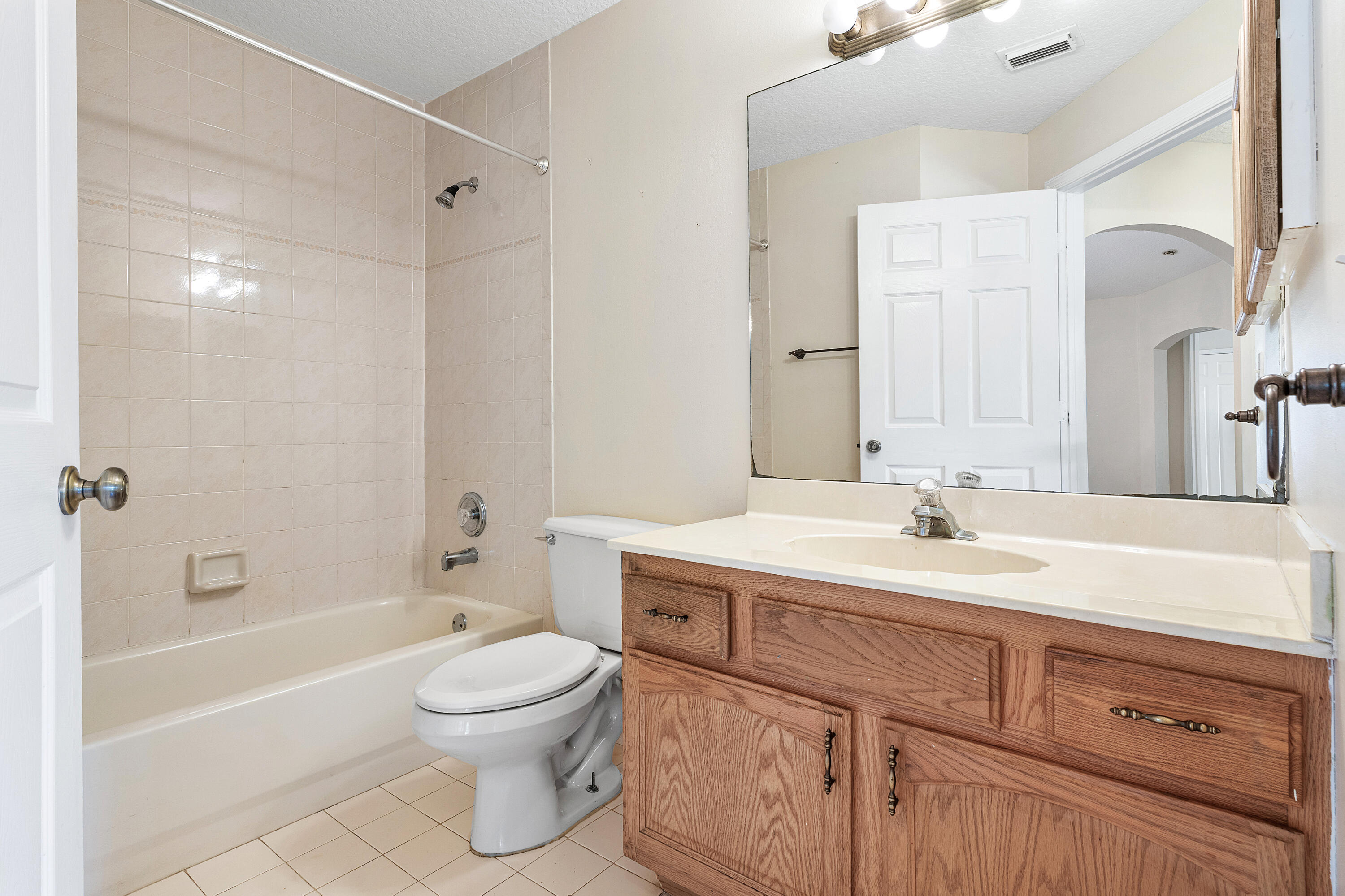 Guest bathroom featuring a wood-finish vanity with an integrated countertop, large wall-mounted mirror, ceramic tile flooring, and a shower-tub combination with tile surround