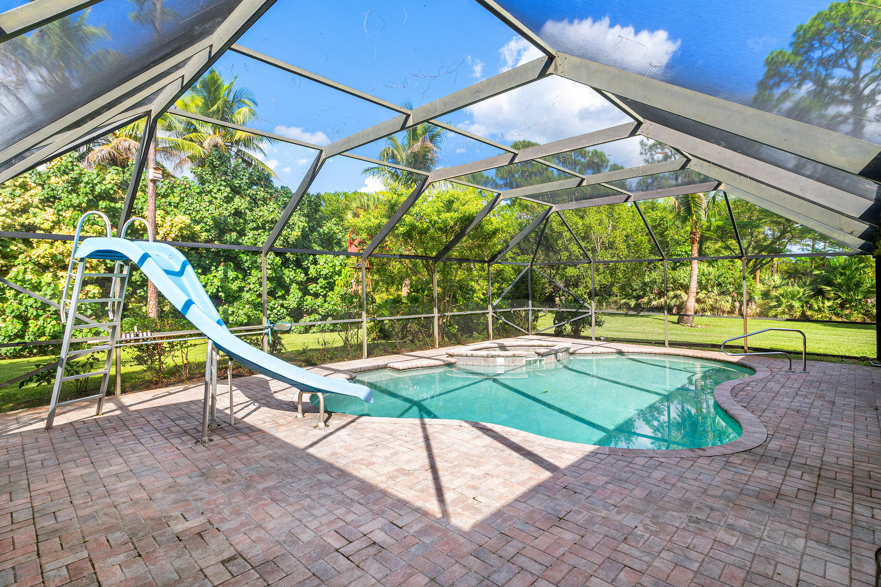 Screened-in pool enclosure featuring paver decking, an integrated pool slide, and an in-ground spa