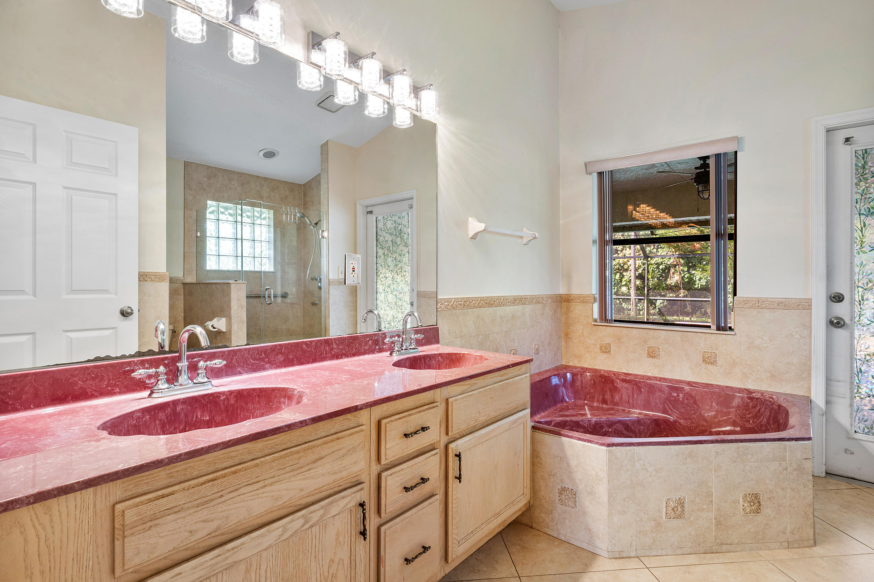 Primary bathroom ensuite number one double vanity featuring a wide countertop with dual integrated sinks, light wood cabinetry, a large wall-mounted mirror, multi-bulb overhead lighting, and a corner soaking tub finished with decorative stone tile accents