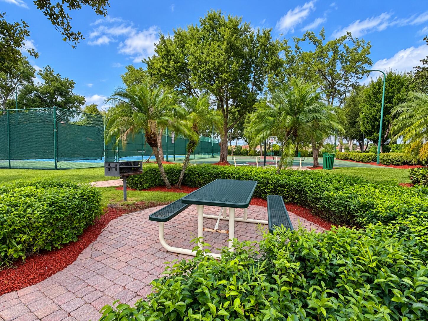 Outdoor recreation area featuring a paved brick patio, built-in charcoal grill, tennis court, and manicured landscaping with mature trees and palms