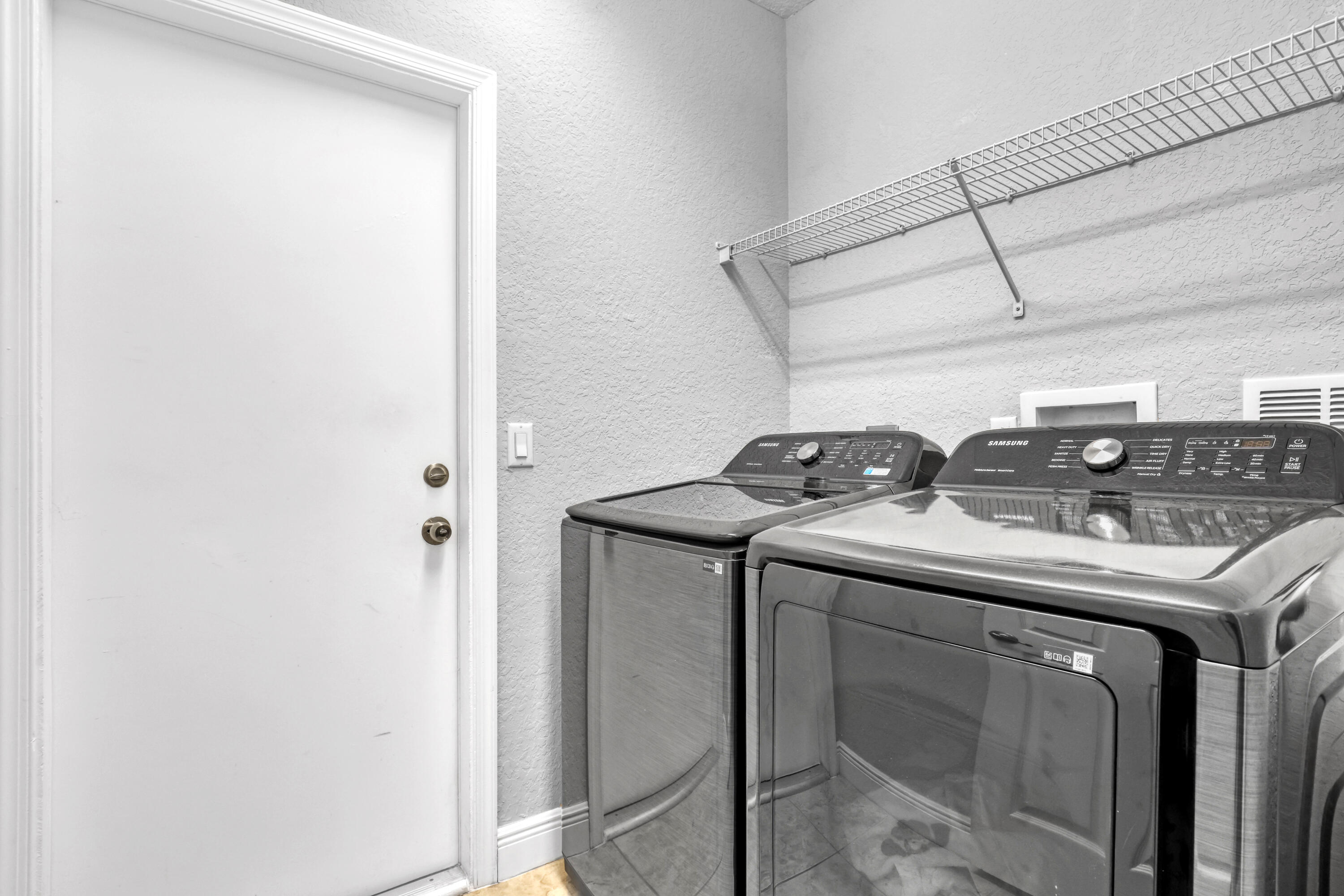 Laundry space featuring metallic-finish top-load washer and dryer with glass lids, paired with an integrated wall-mounted wire shelving unit and a solid-core entry door to garage