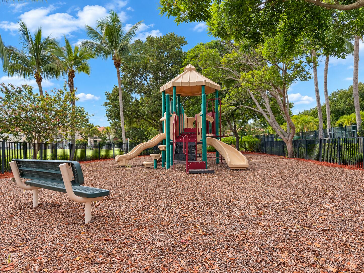 Outdoor recreation area featuring a multi-level play structure with dual slides, wood-chip ground cover, a metal-frame bench, and perimeter fencing shaded by mature palm and deciduous trees