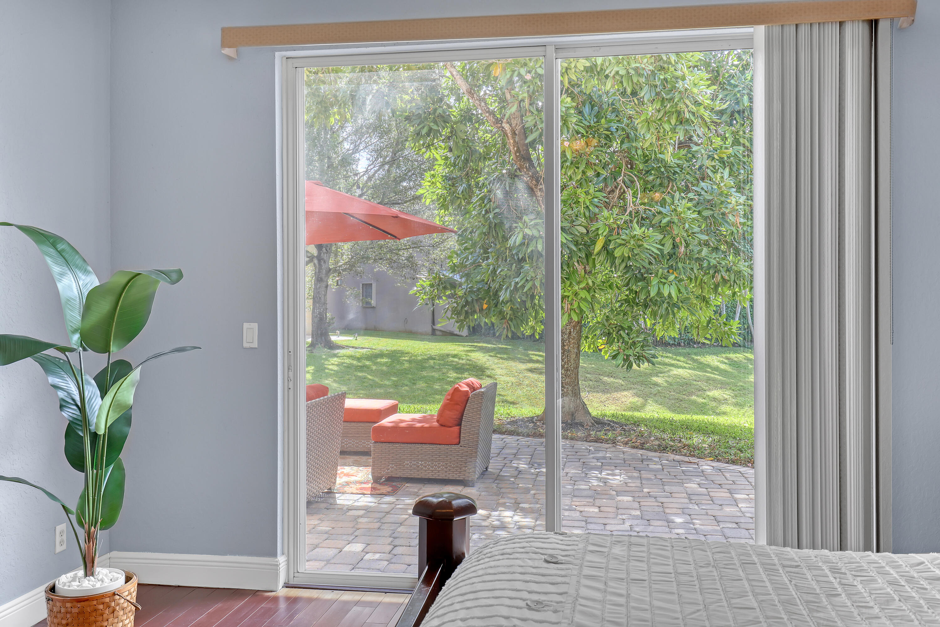 Sliding glass door with vertical blinds opening to a paved stone patio, mature trees, and a grass lawn