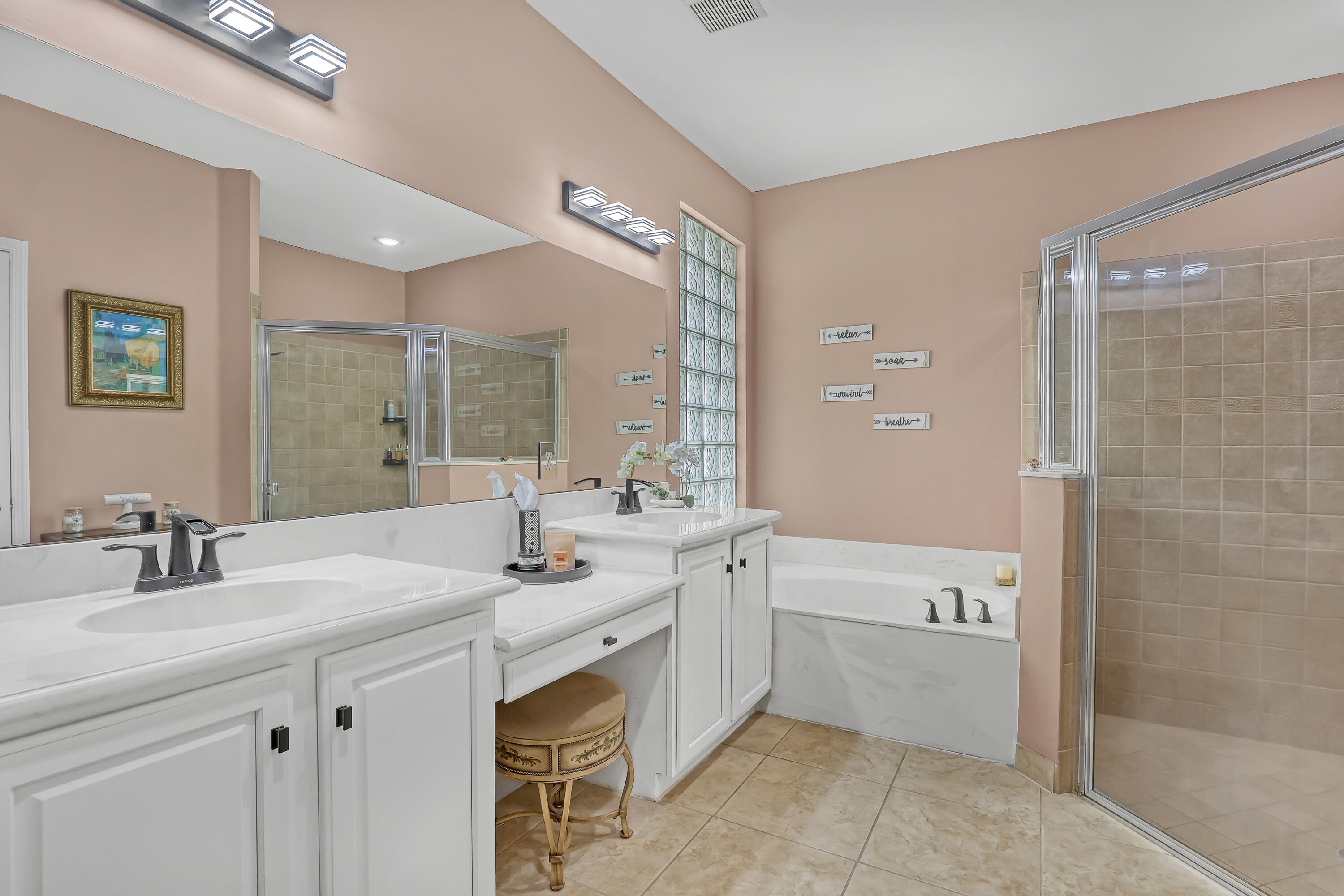 Dual-sink vanity with white cabinetry and stone countertops, glass block window, soaking tub, and a walk-in shower with glass enclosure